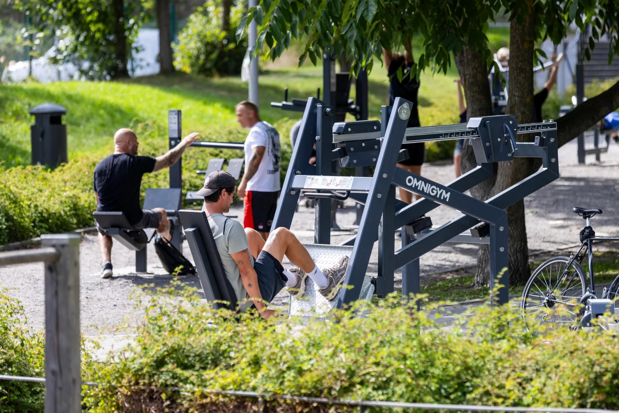 Utomhusgym med människor som tränar på olika träningsmaskiner i en grön parkmiljö.