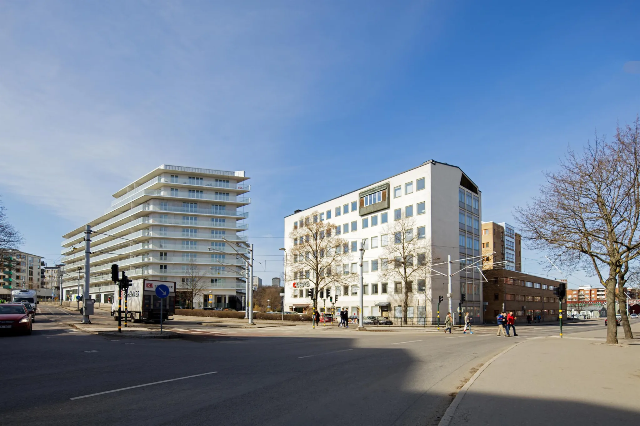 En korsning med moderna byggnader, inklusive ett vitt kontorshus och ett glasfasadhus, under en klarblå himmel.