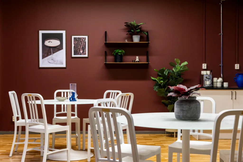 Ett caféområde med vita bord och stolar, växter och tavlor på en brun vägg.