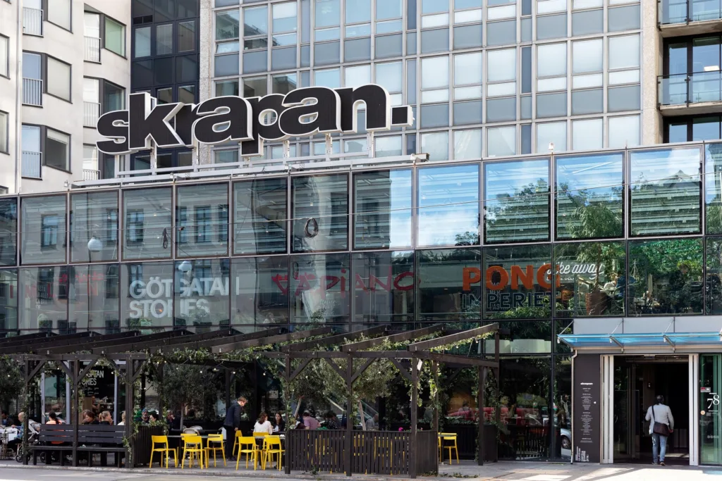 1. En modern byggnad med glasfasad och texten "Kapan" synlig på toppen. Nedanför finns en restaurang med uteservering och gula stolar.