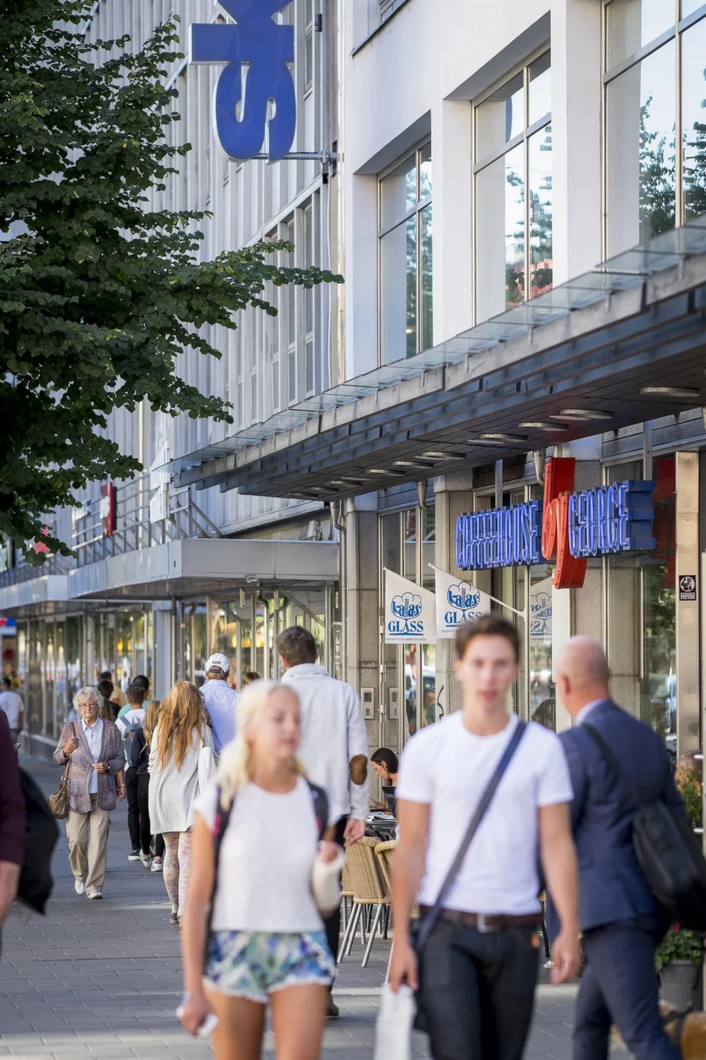 En livlig gatuscen med människor som promenerar framför butiker och skyltfönster längs trottoaren.