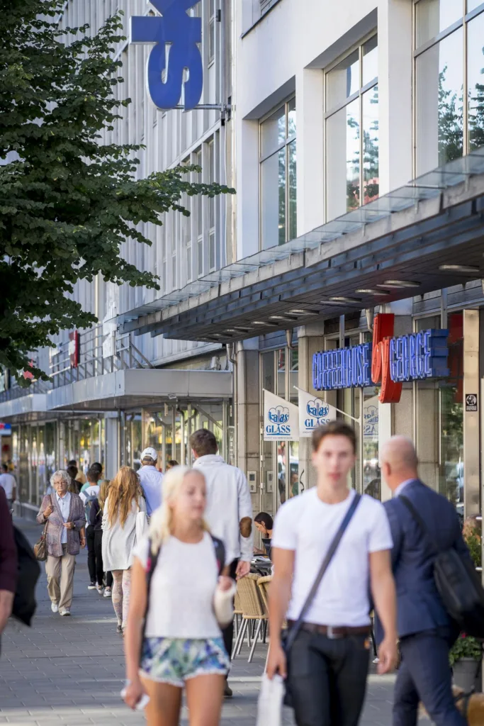 En livlig gatuscen med människor som promenerar framför butiker och skyltfönster längs trottoaren.