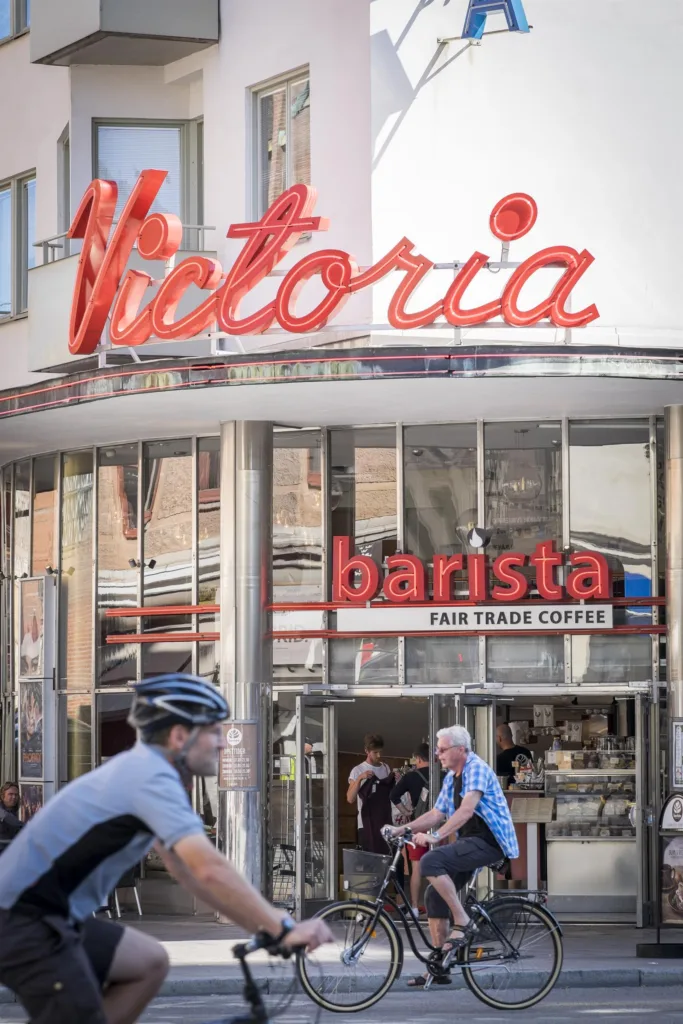 2. En fasad med neonskylten "Victoria" ovanför entrén till ett café med texten "barista fair trade coffee", cyklister syns i förgrunden.