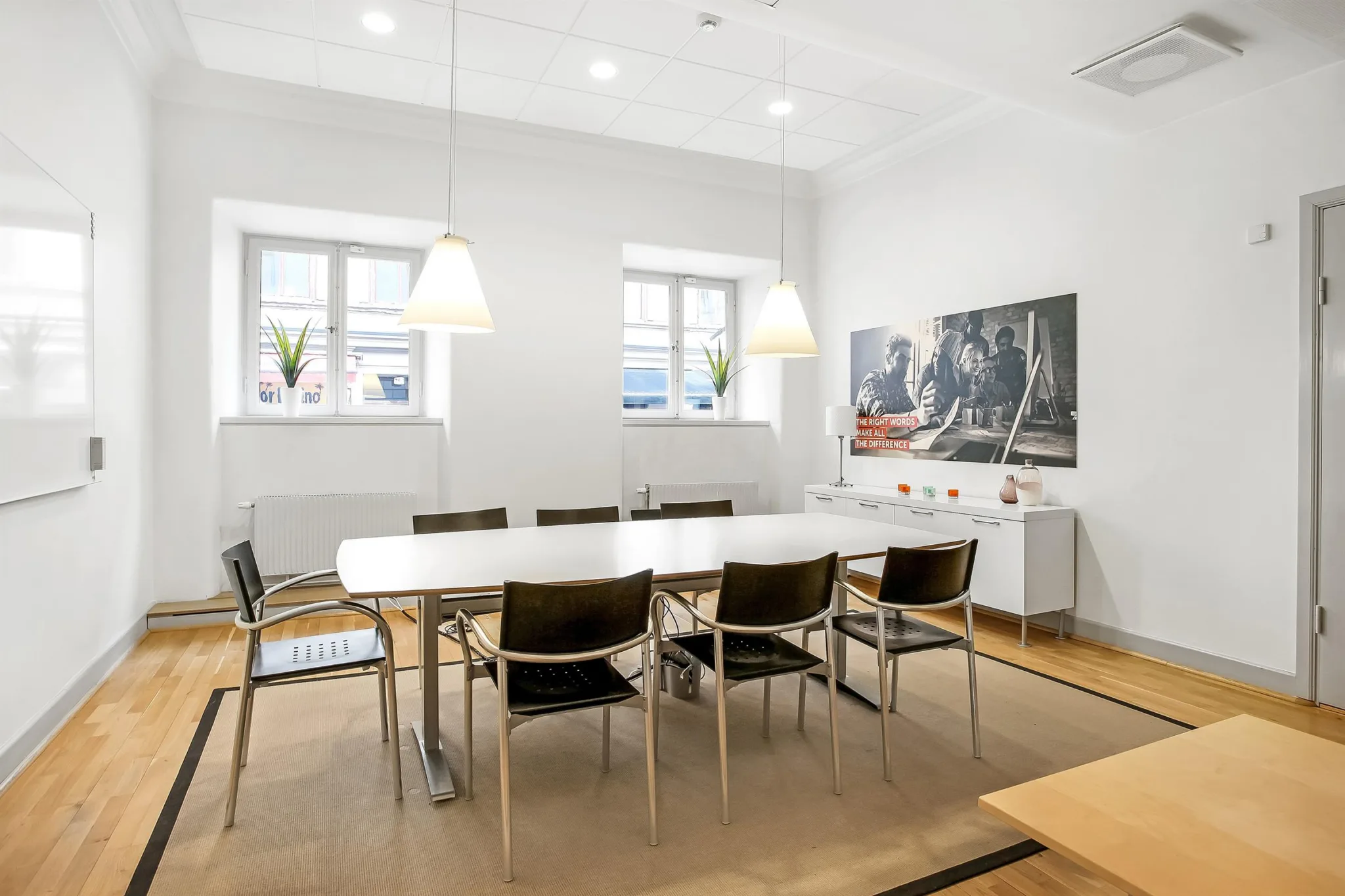 Ett konferensrum med ett vitt bord, svarta stolar, tavlor på väggen och två fönster.