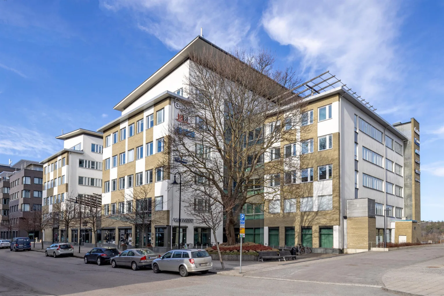 1. En modern kontorsbyggnad i flera våningar omgiven av parkerade bilar och träd under en klarblå himmel.