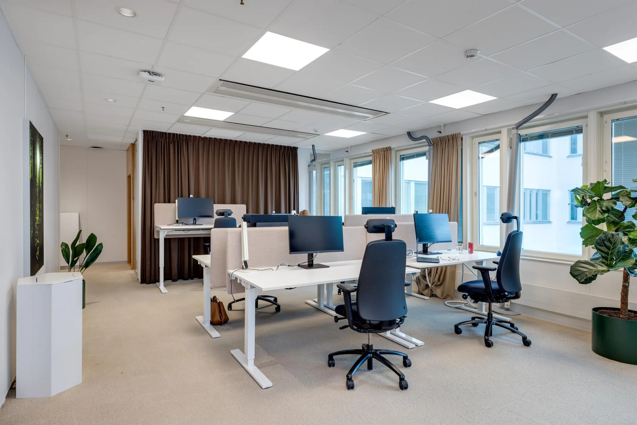 1. Ett öppet kontorslandskap med höj- och sänkbara skrivbord, kontorsstolar och datorskärmar, omgiven av stora fönster och gardiner.