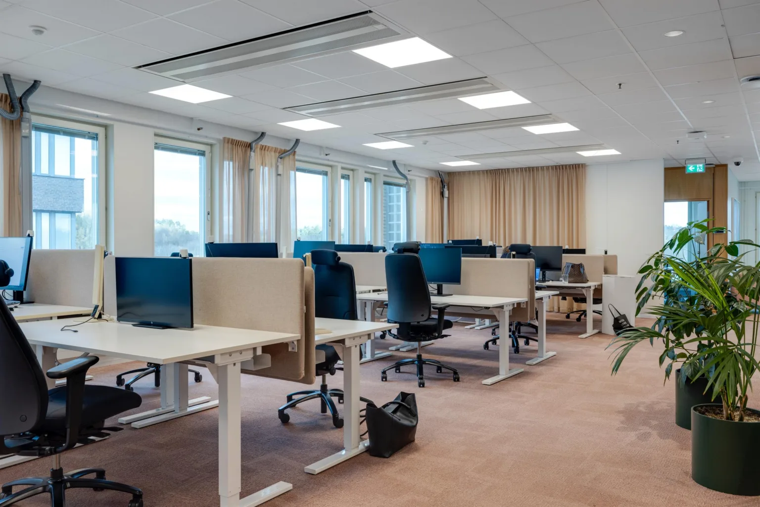 1. Ett kontorsutrymme med skrivbord, kontorsstolar och datorer, omgivna av stora fönster och gardiner.