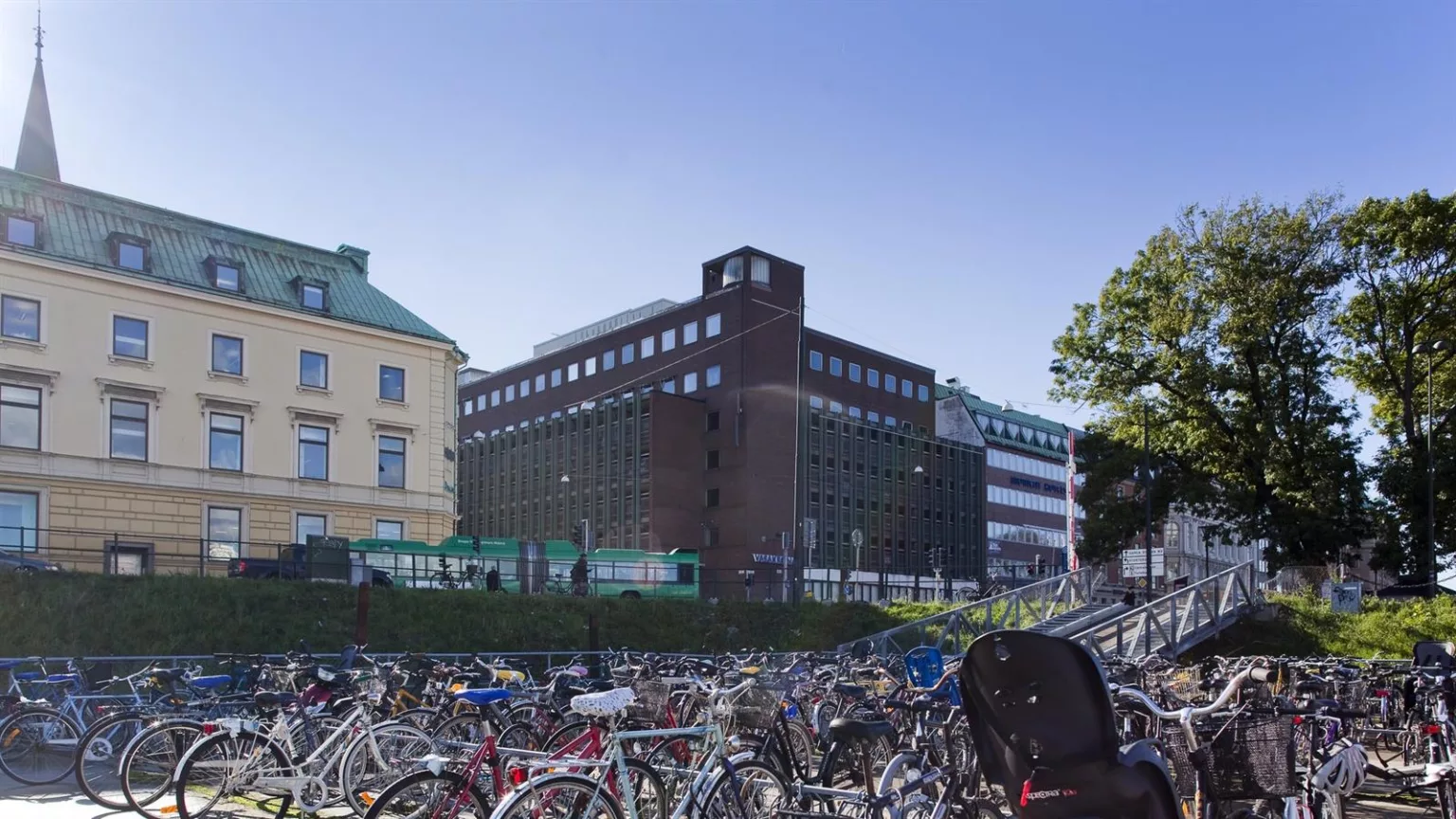 - En stadsmiljö med ett mörkt tegelhus i bakgrunden, omgiven av cykelställ fyllda med cyklar.