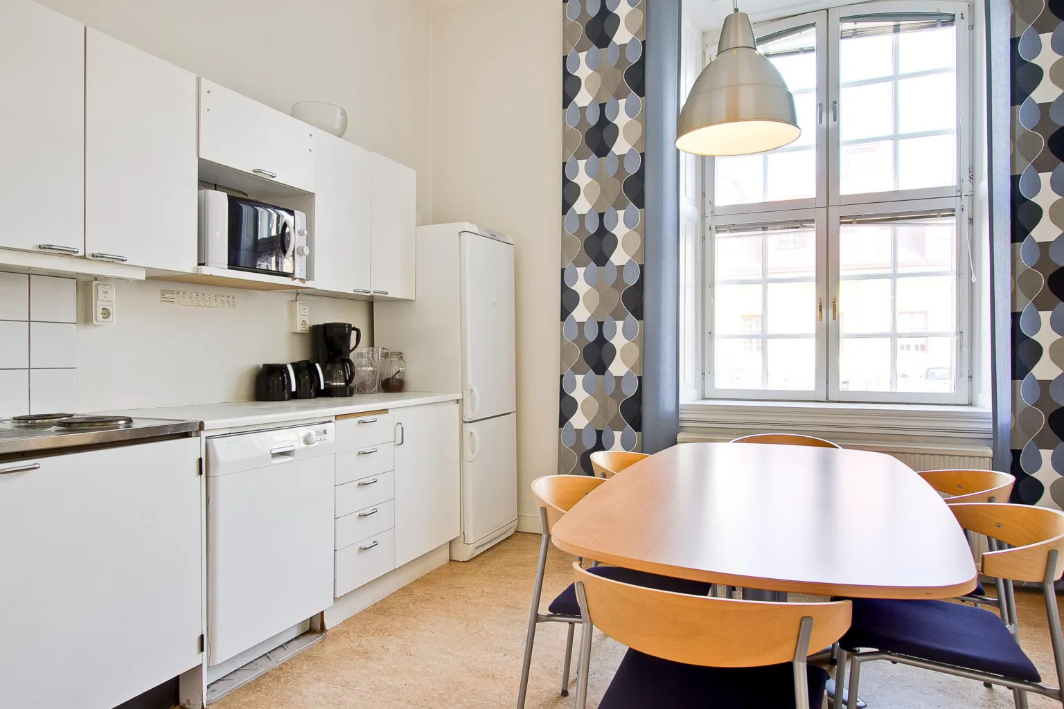 3. Ett kök med vita skåp, en mikrovågsugn och kaffebryggare, ett ovalt träbord med fyra stolar och geometriska gardiner vid fönstret.