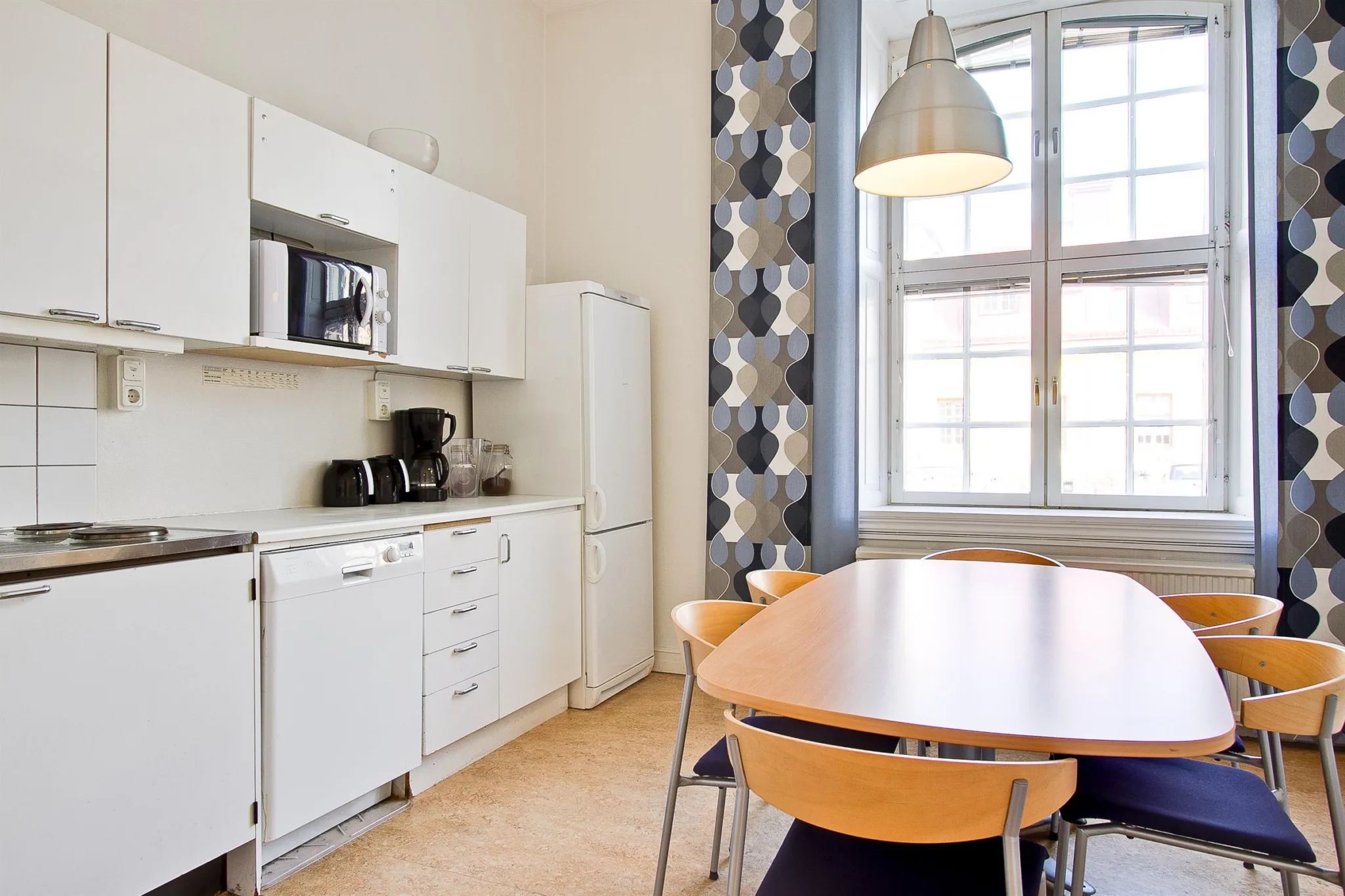 3. Ett kök med vita skåp, en mikrovågsugn och kaffebryggare, ett ovalt träbord med fyra stolar och geometriska gardiner vid fönstret.