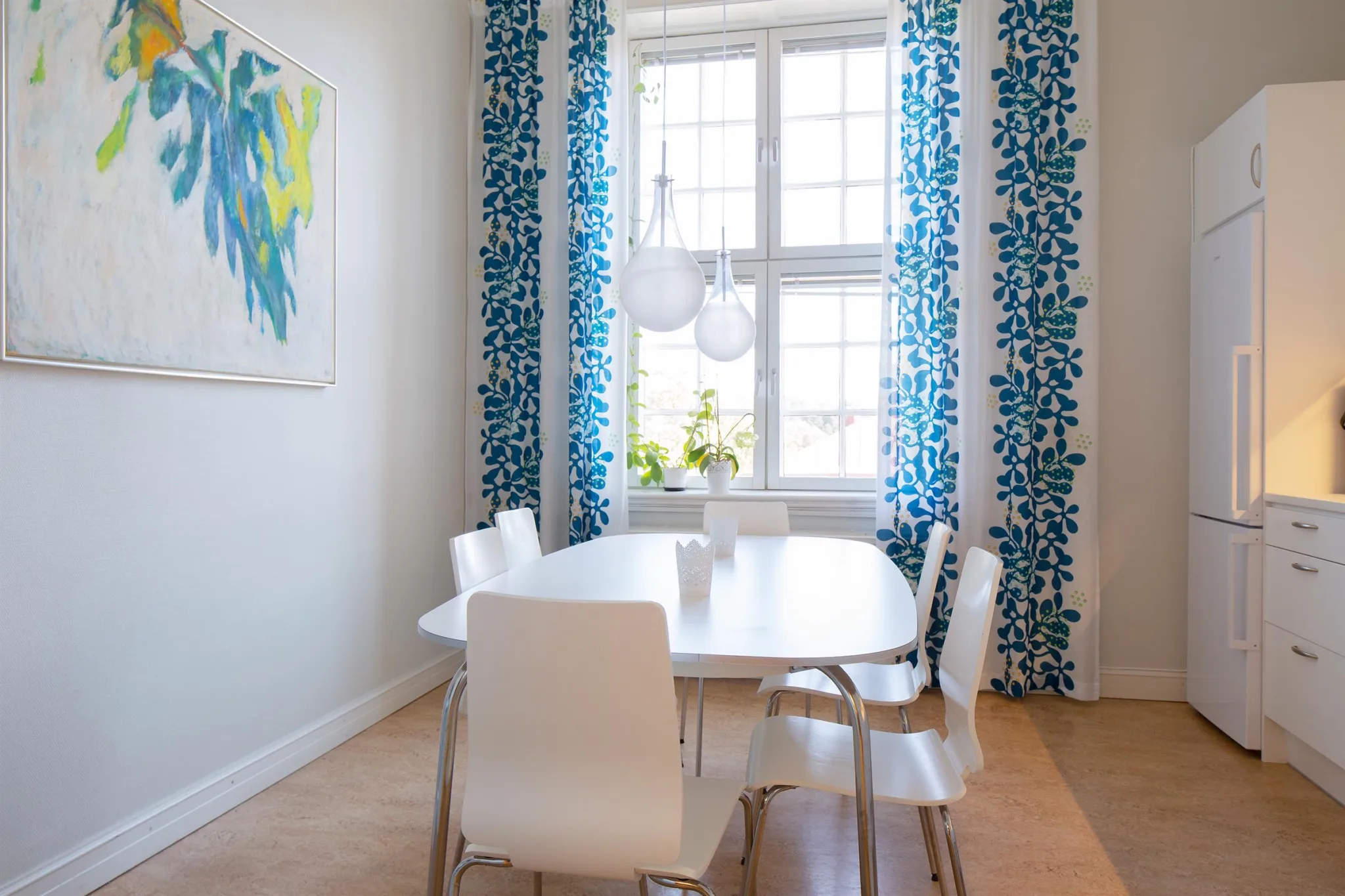 2. Ett matsalsrum med ett vitt bord och sex vita stolar, blåmönstrade gardiner och ett stort fönster i bakgrunden.