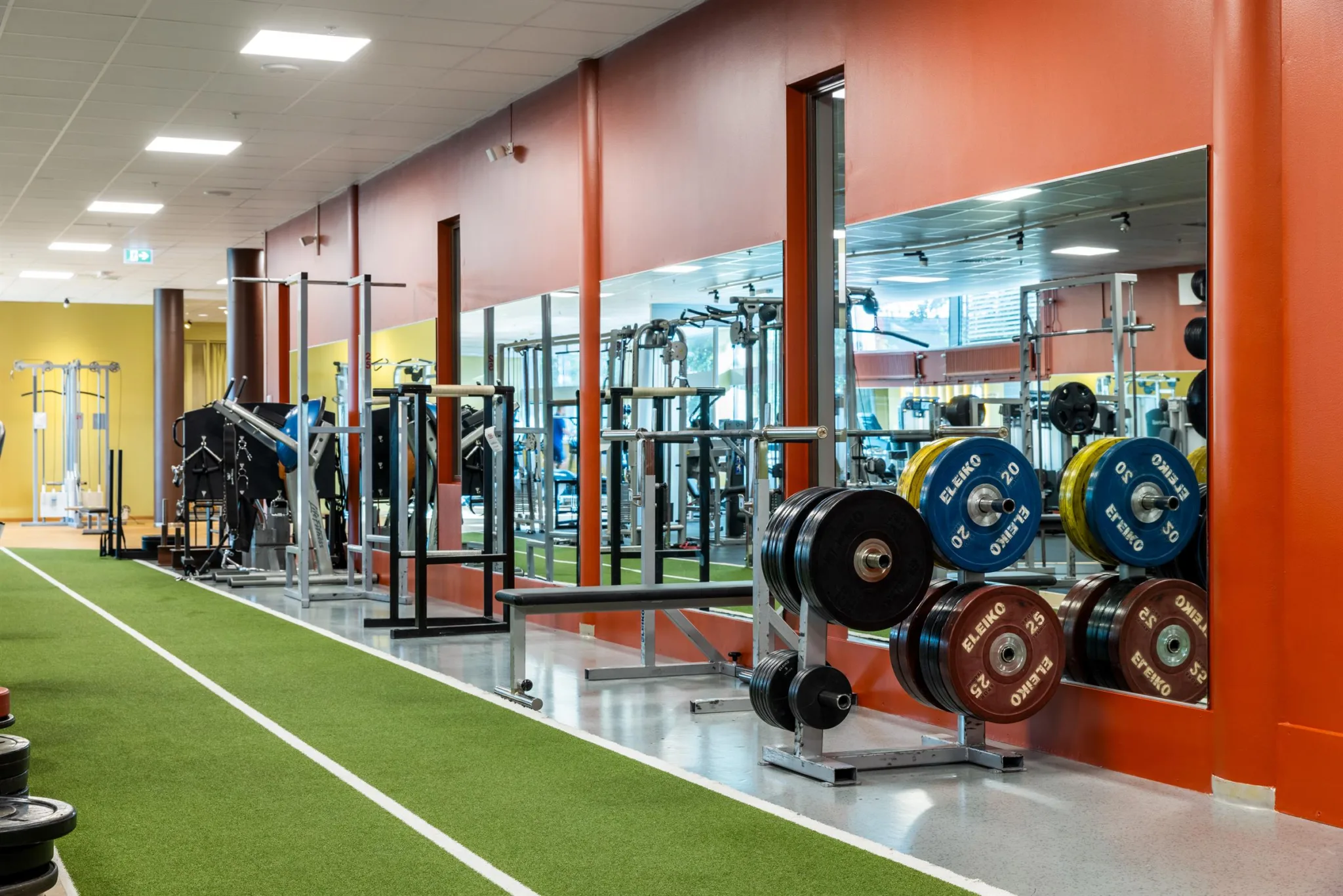 Gym med grön löparbana, vikter och spegelväggar med reflektioner av träningsmaskiner.