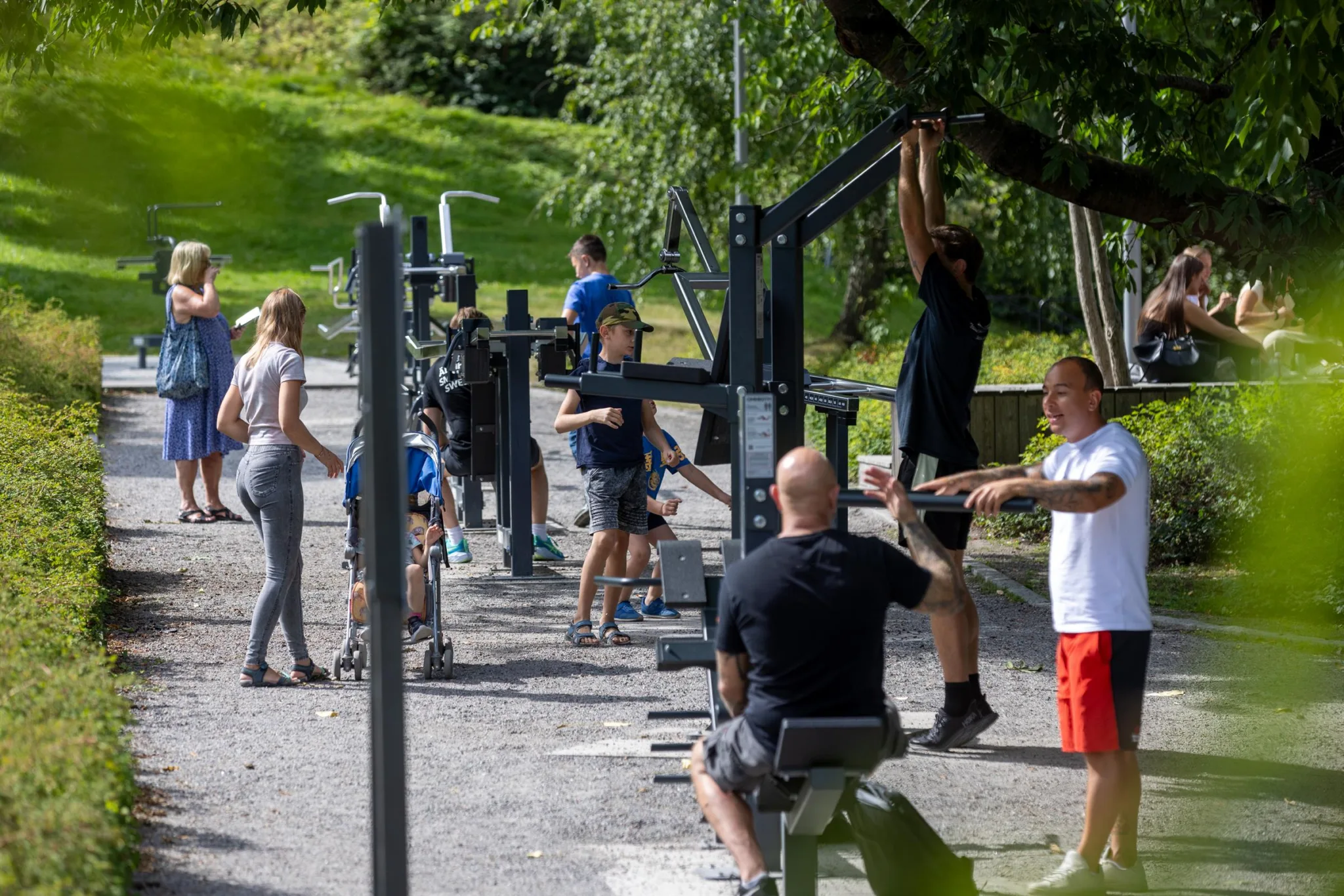 En utomhusgymplats med flera personer som använder träningsmaskiner, omgiven av grönska.