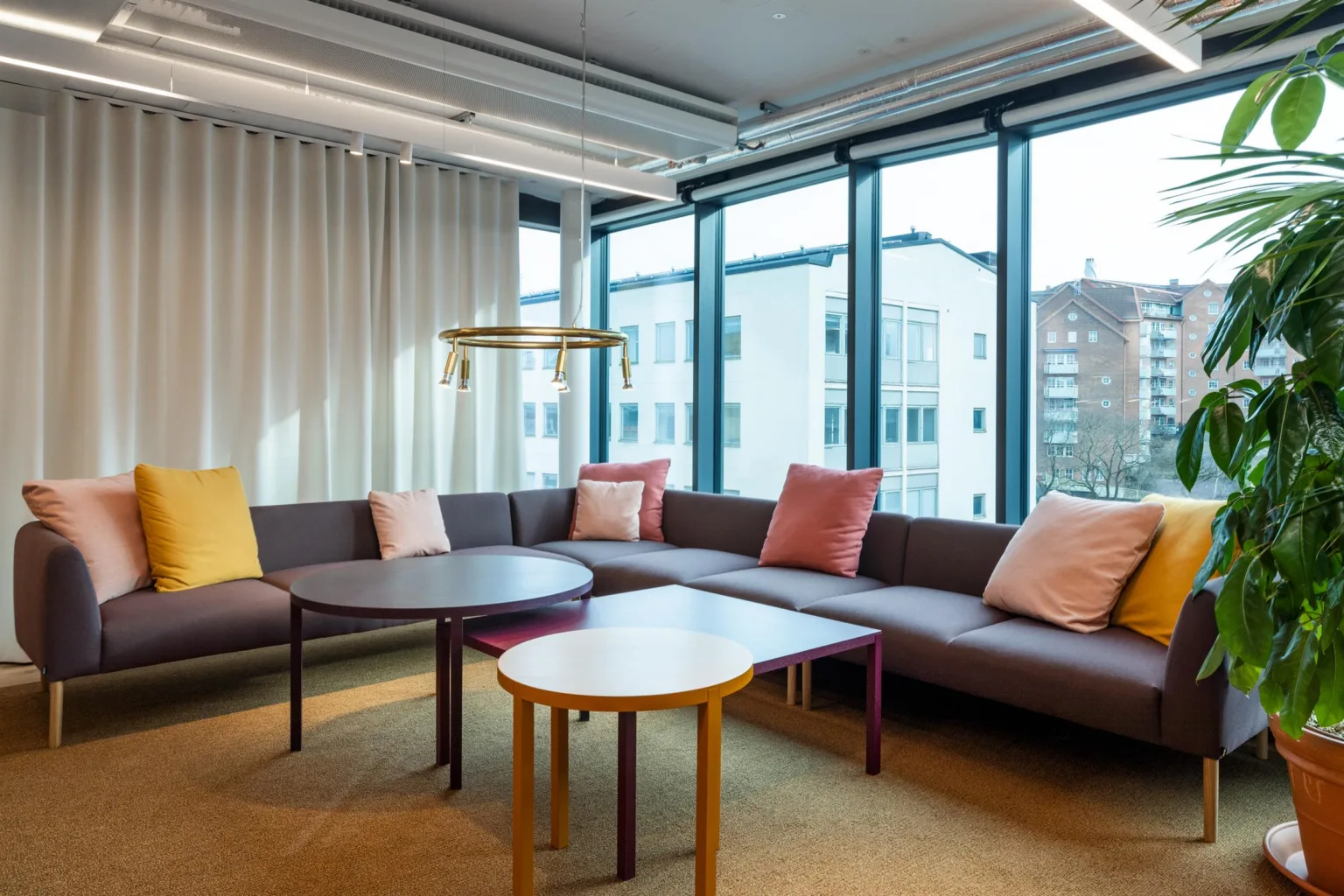 1. En modern lounge med grå soffa, färgglada kuddar, runda bord och stora fönster som släpper in dagsljus.