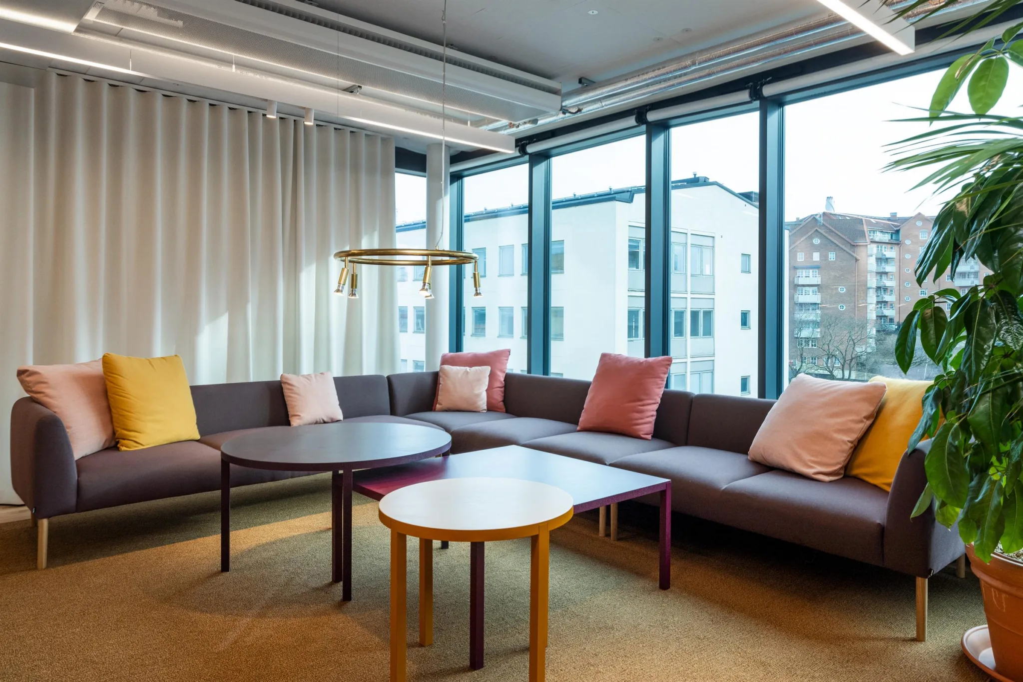 1. En modern lounge med grå soffa, färgglada kuddar, runda bord och stora fönster som släpper in dagsljus.