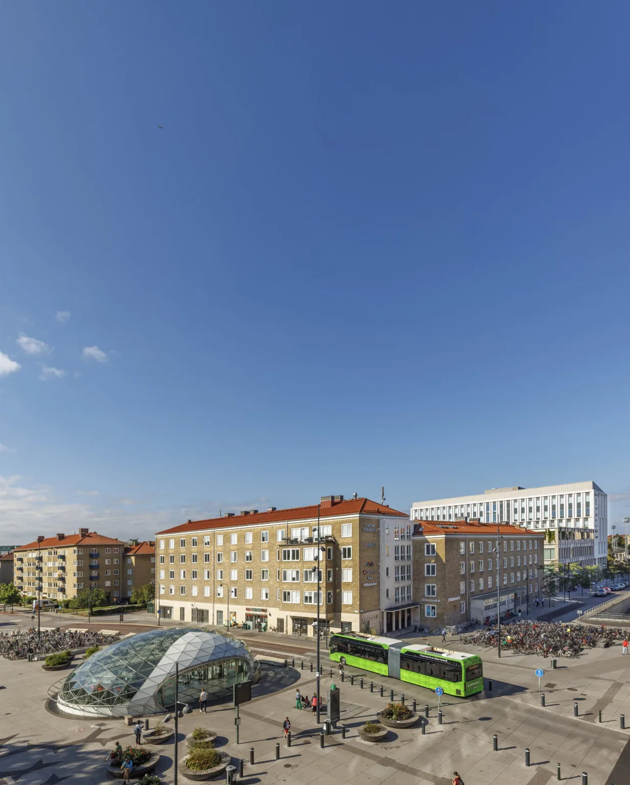 En stadsvy med blå himmel, äldre byggnader med röda tak och en grön spårvagn.