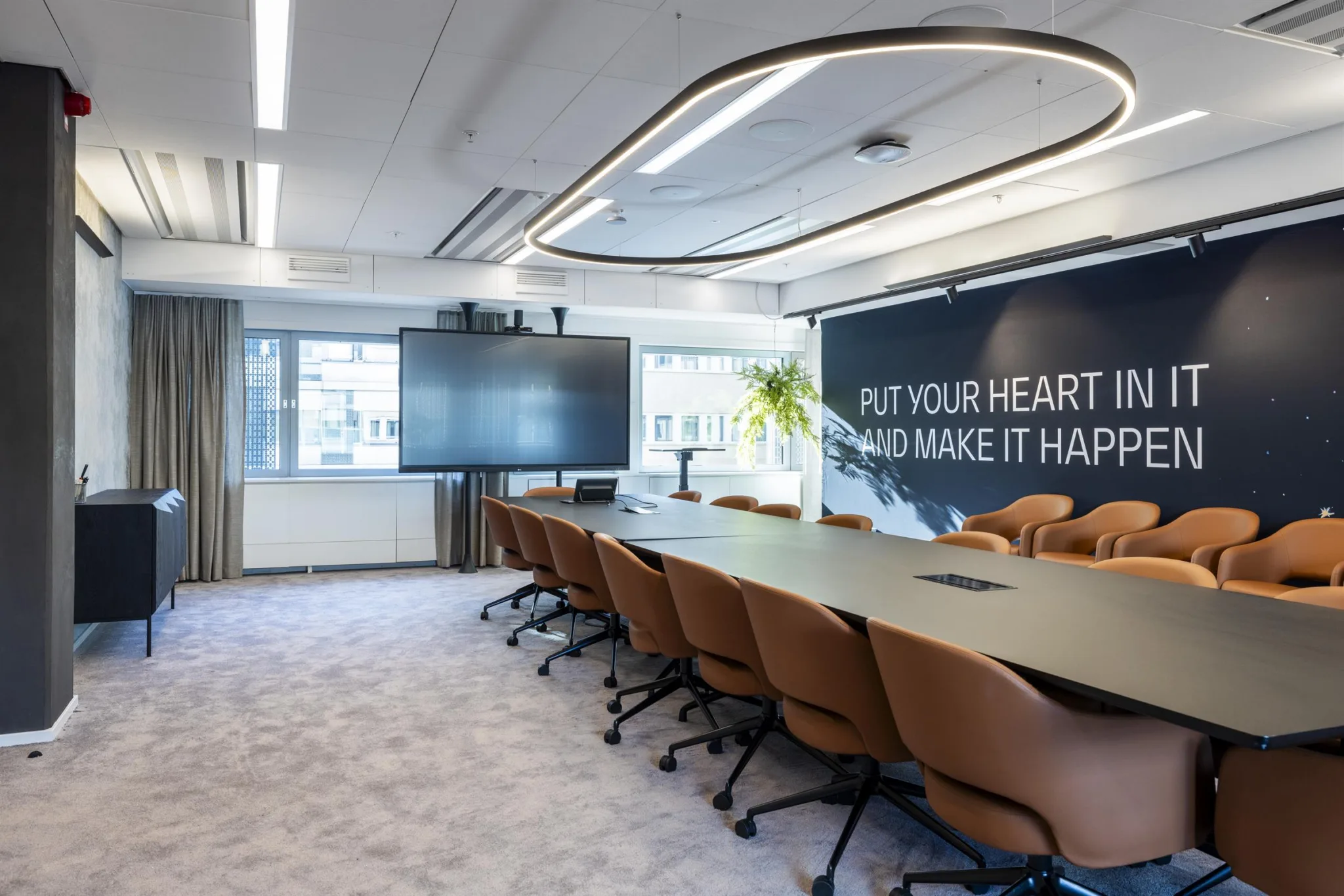 1. Ett modernt konferensrum med ett långt bord och bruna stolar, en stor skärm och texten "Put your heart into it and make it happen" på väggen.