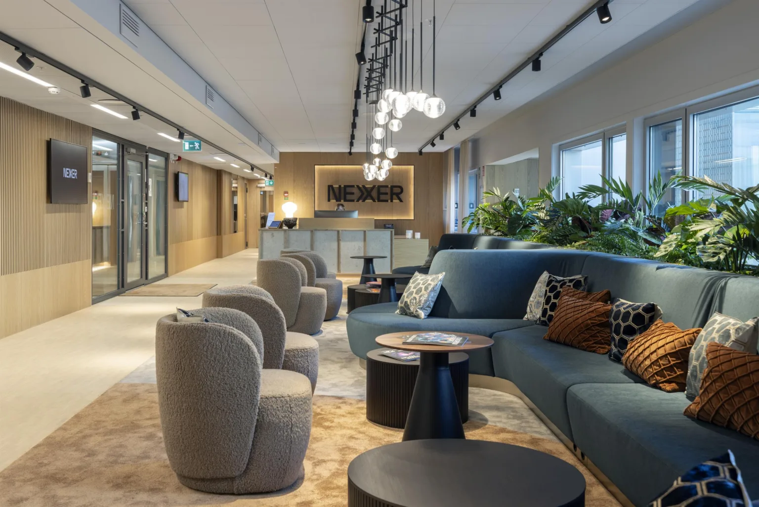 2. Ett stilrent loungeområde med soffor, fåtöljer, runda bord och växter, samt belysning i taket och en reception i bakgrunden.