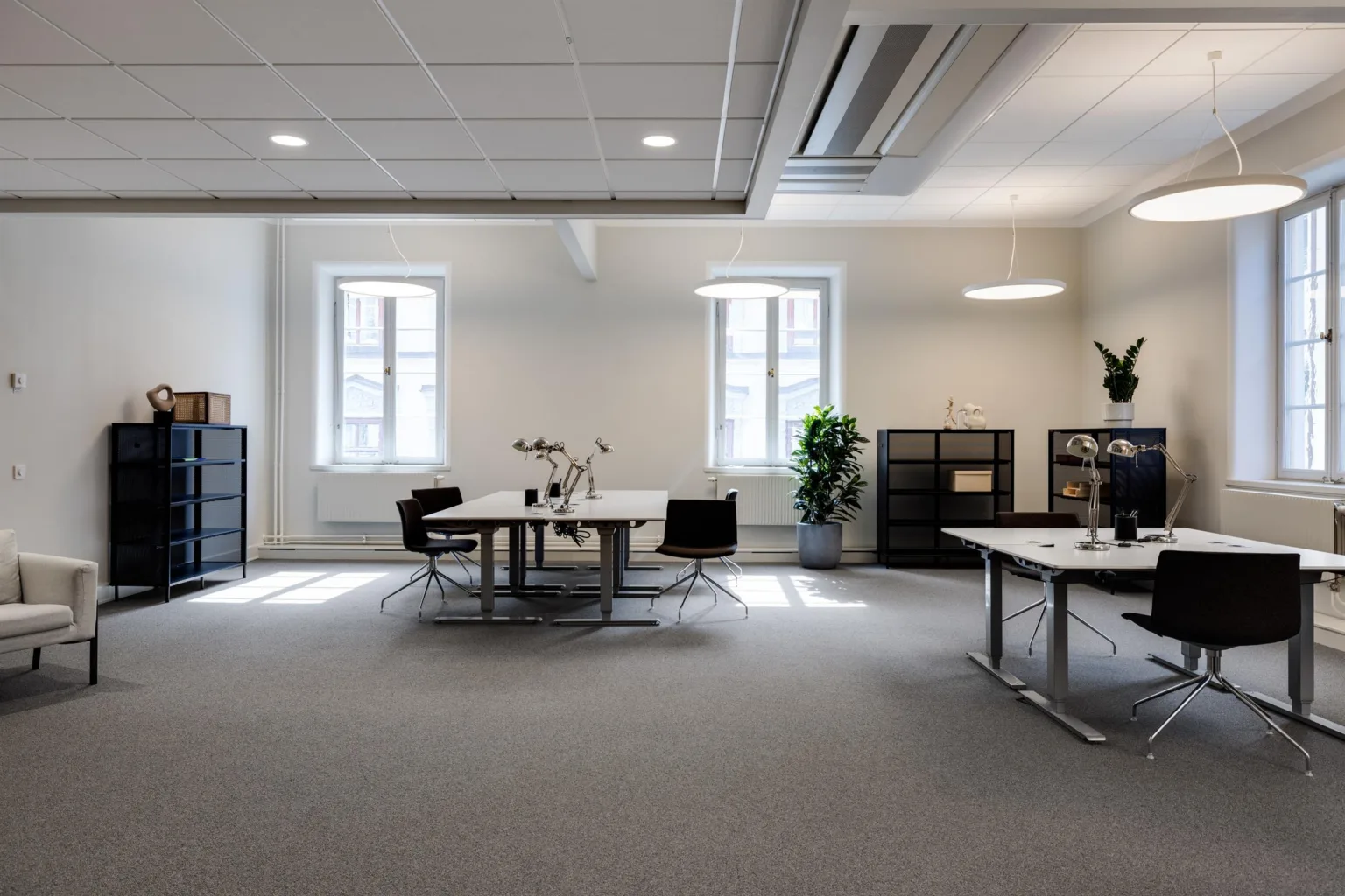 Ett rymligt kontor med flera arbetsstationer, svarta stolar, hyllor och växter under takbelysning.