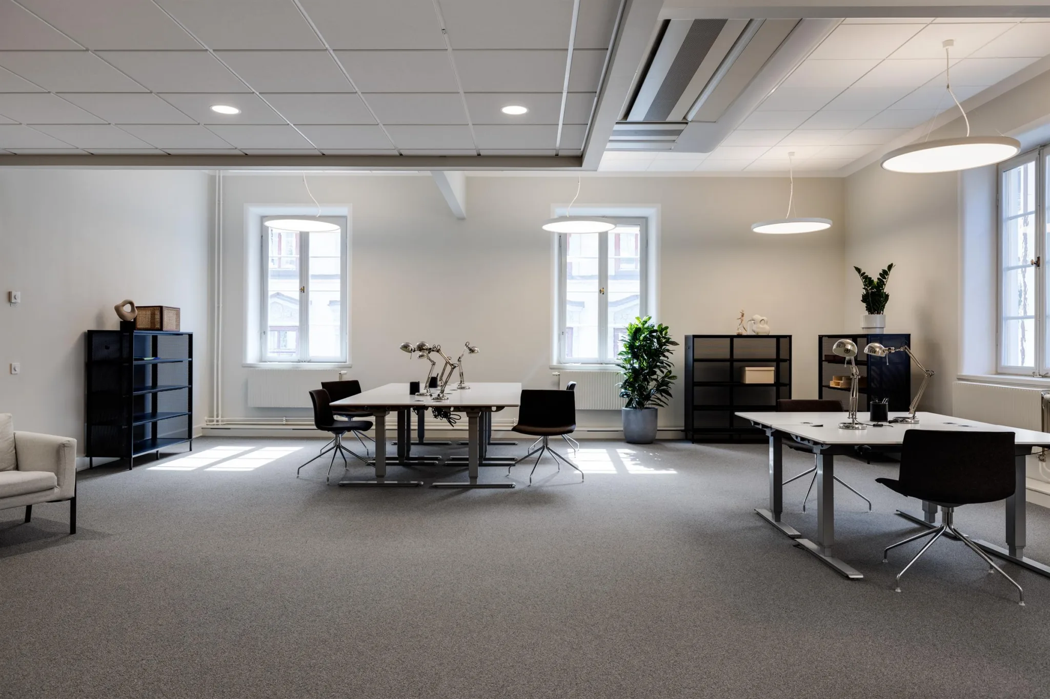 Ett rymligt kontor med flera arbetsstationer, svarta stolar, hyllor och växter under takbelysning.