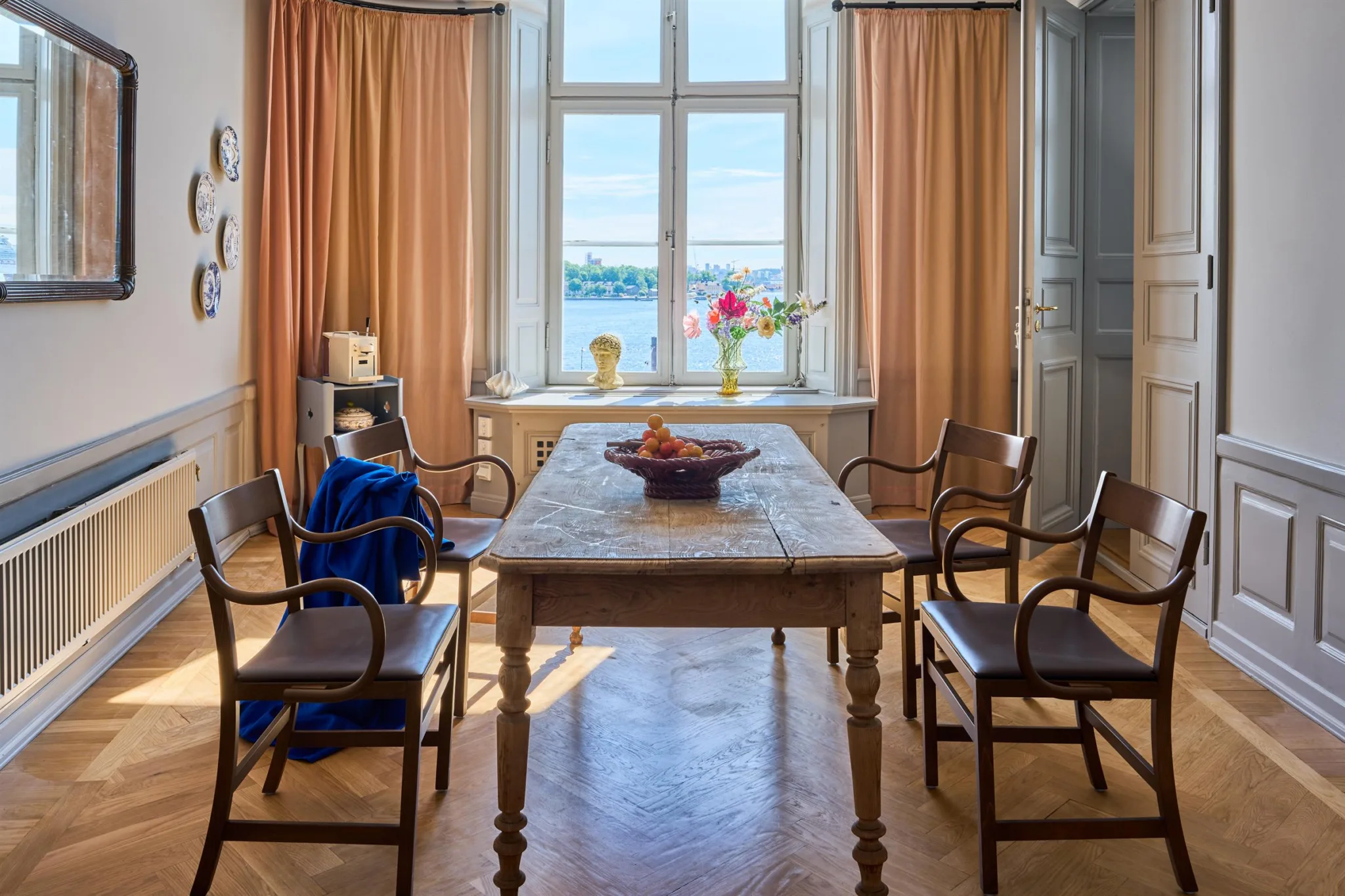 Ett rum med ett stort träbord, fyra stolar och orange gardiner, med utsikt över vatten genom ett fönster.
