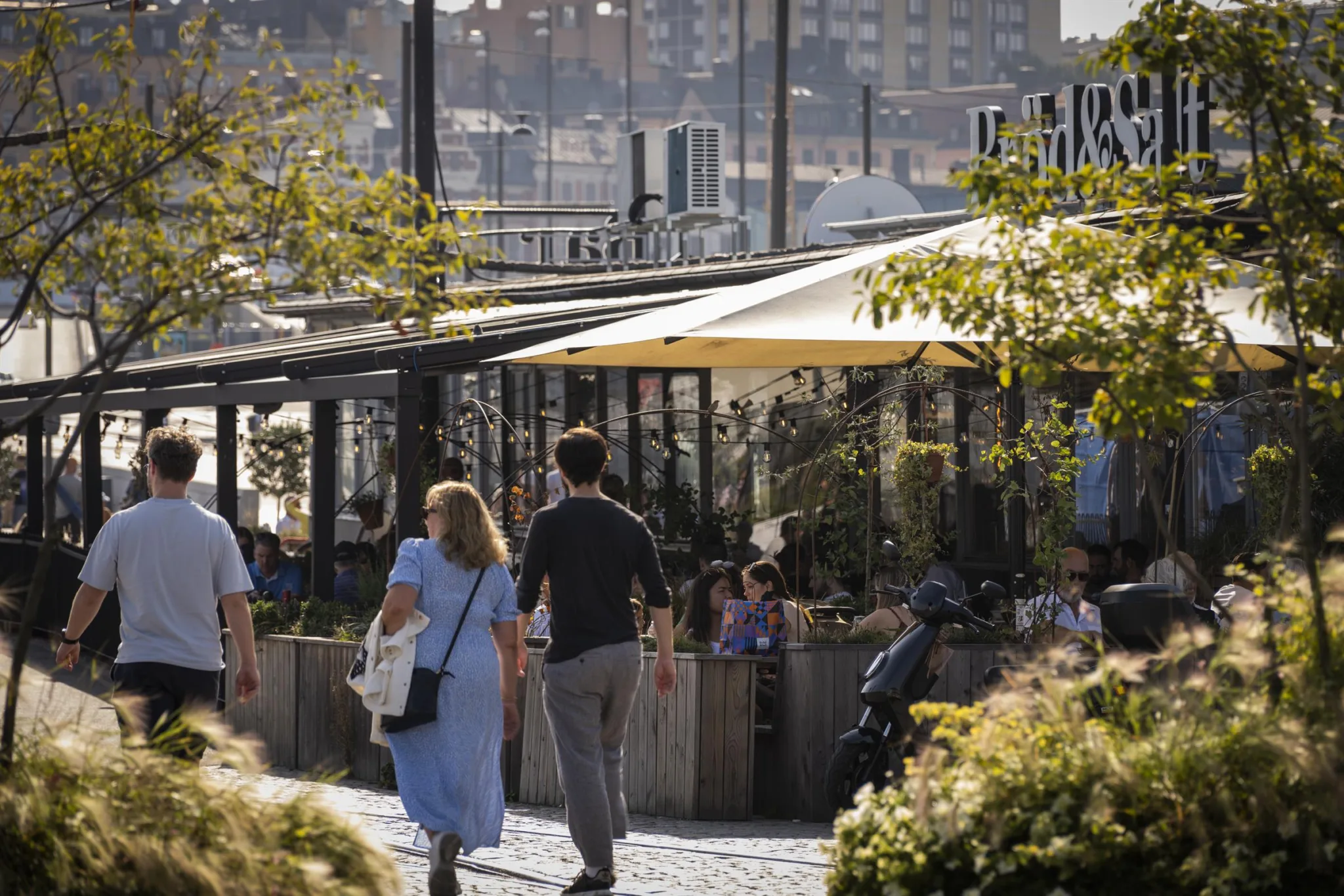 Människor promenerar vid en uteservering med växter och parasoller, med stadsmiljö i bakgrunden.