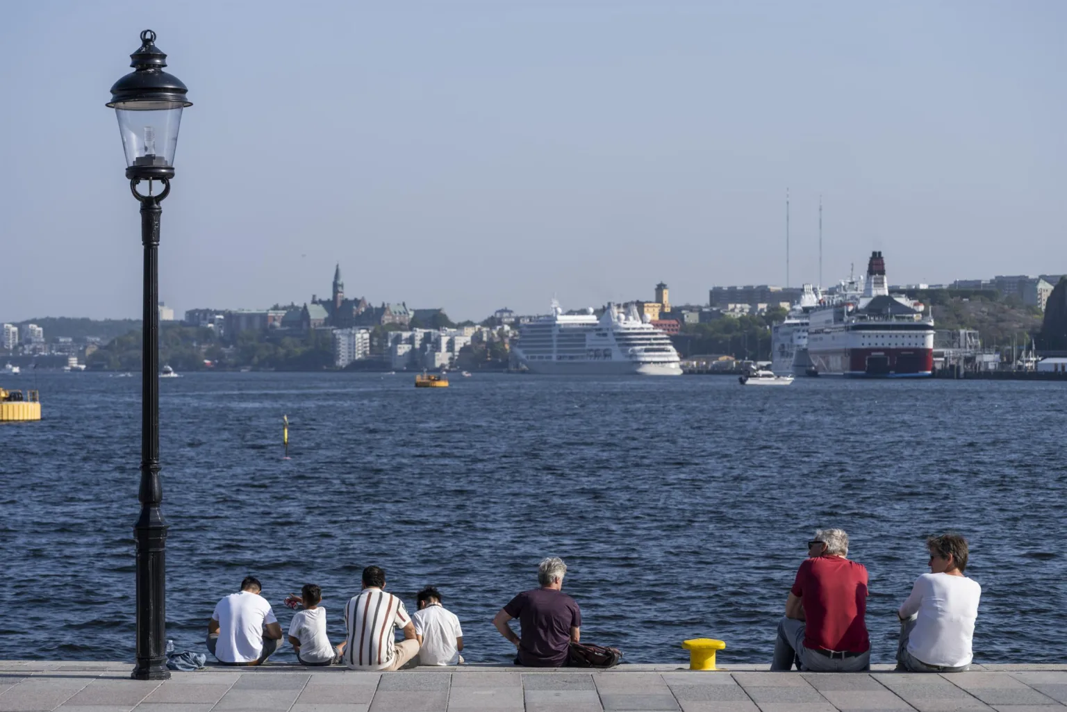 En grupp människor sitter vid en kaj med havet och kryssningsfartyg i bakgrunden.