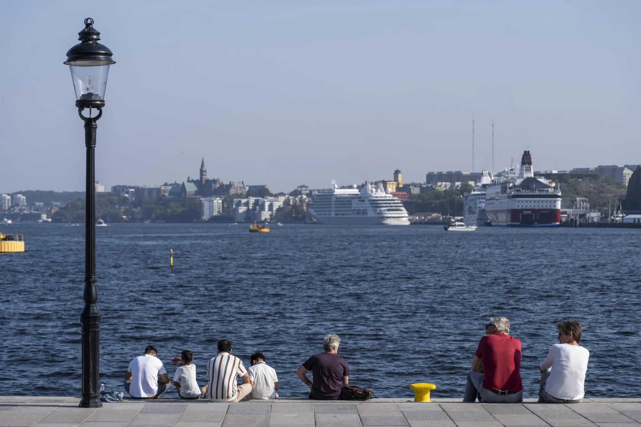 En grupp människor sitter vid en kaj med havet och kryssningsfartyg i bakgrunden.