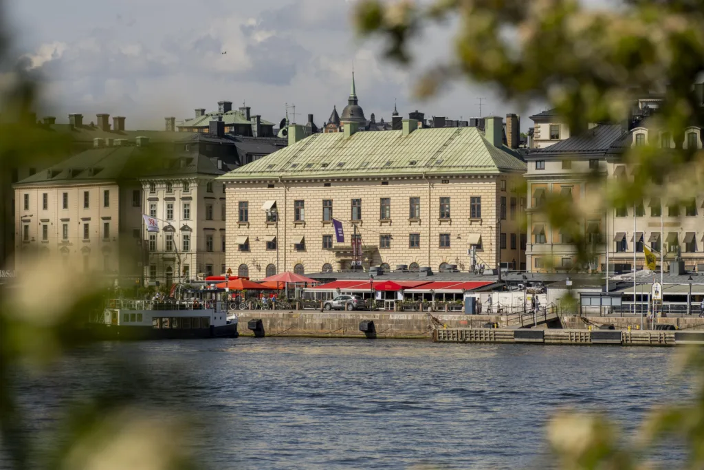 En byggnad vid vattnet med grön takbeklädnad, röda markiser och omgivande stadsmiljö.