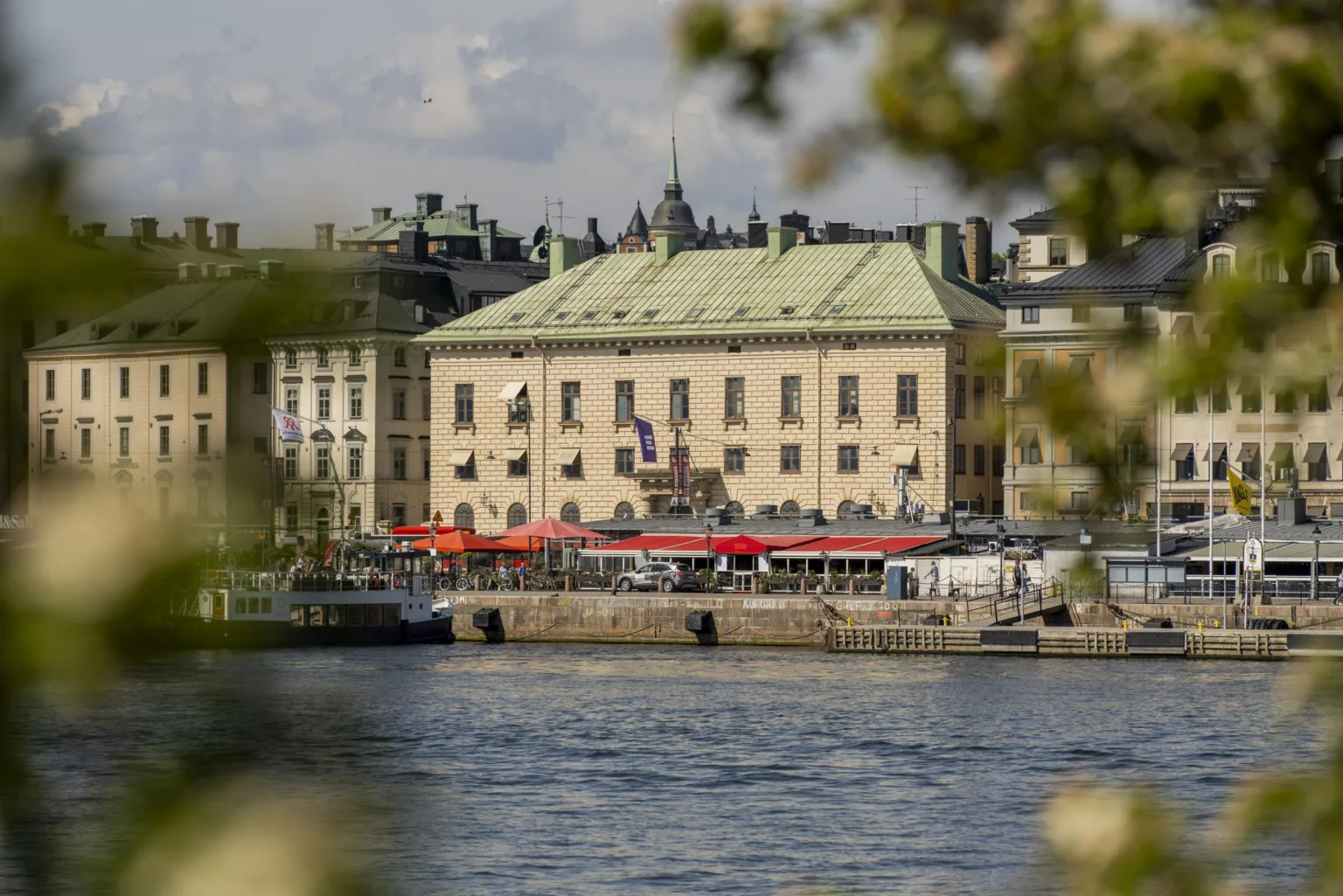 En byggnad vid vattnet med grön takbeklädnad, röda markiser och omgivande stadsmiljö.