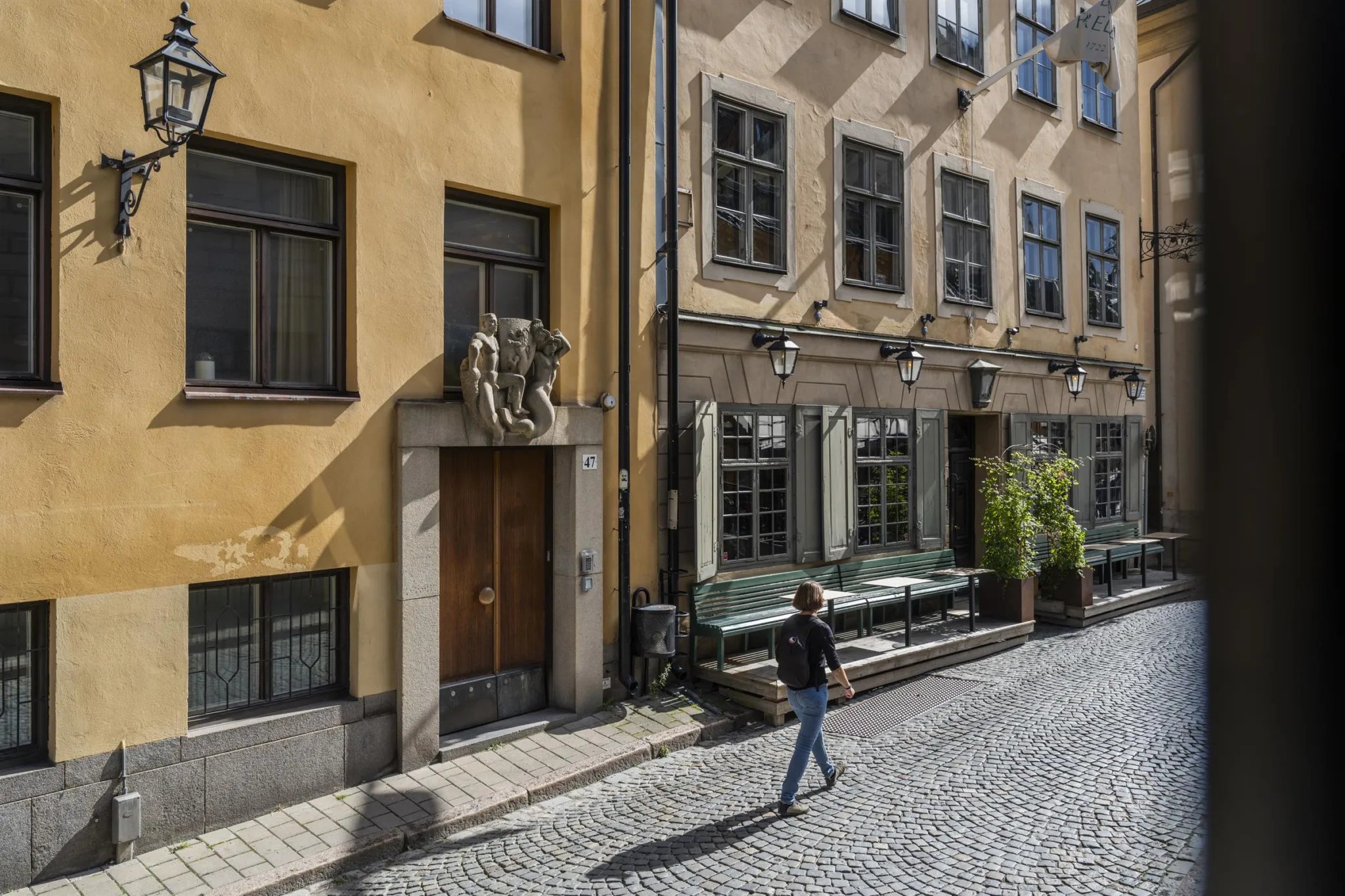 En kullerstensgata med en gul byggnad, en person som går förbi och en entré med en staty ovanför dörren.