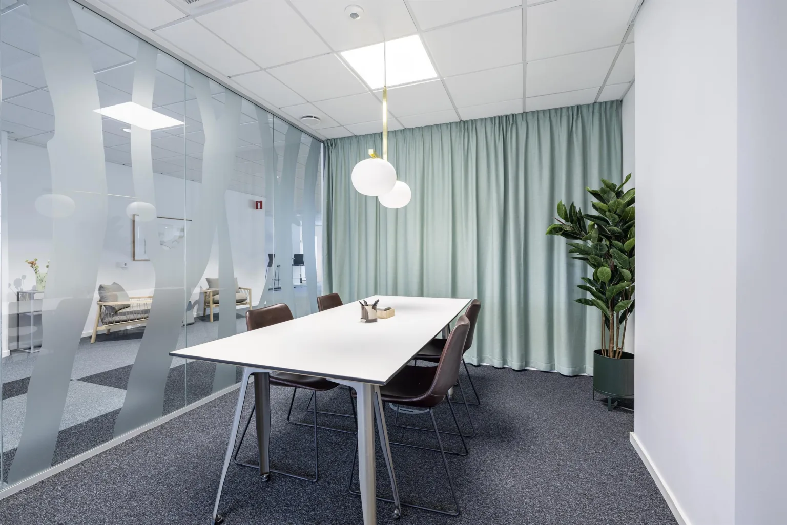 2. Ett identiskt mötesrum med ett vitt bord, fyra stolar, gröna gardiner, en hängande lampa och en grön växt i hörnet.