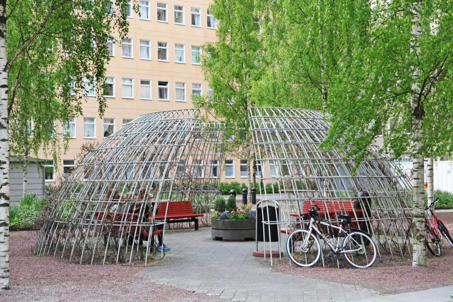 1. En utomhusplats med cykelställ, röda bänkar och ett metallgaller i form av en kupol omgiven av träd och en byggnad i bakgrunden.
