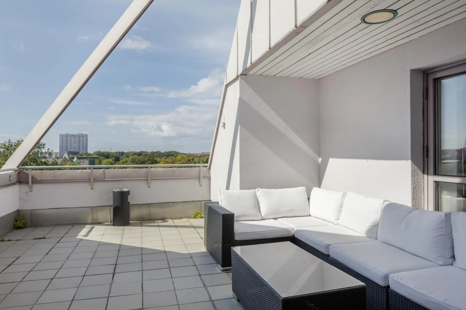 En uteplats med vita soffor och bord under en modern takstruktur, med utsikt över grönska och himmel.