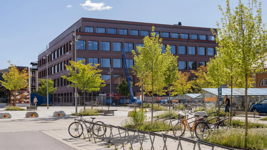 En brun byggnad med stora fönster, framför ett torg med cykelställ och träd.