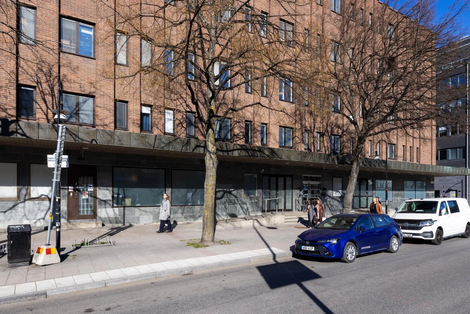 5. Tegelbyggnad med stora fönster och träd framför, en blå bil parkerad på gatan.