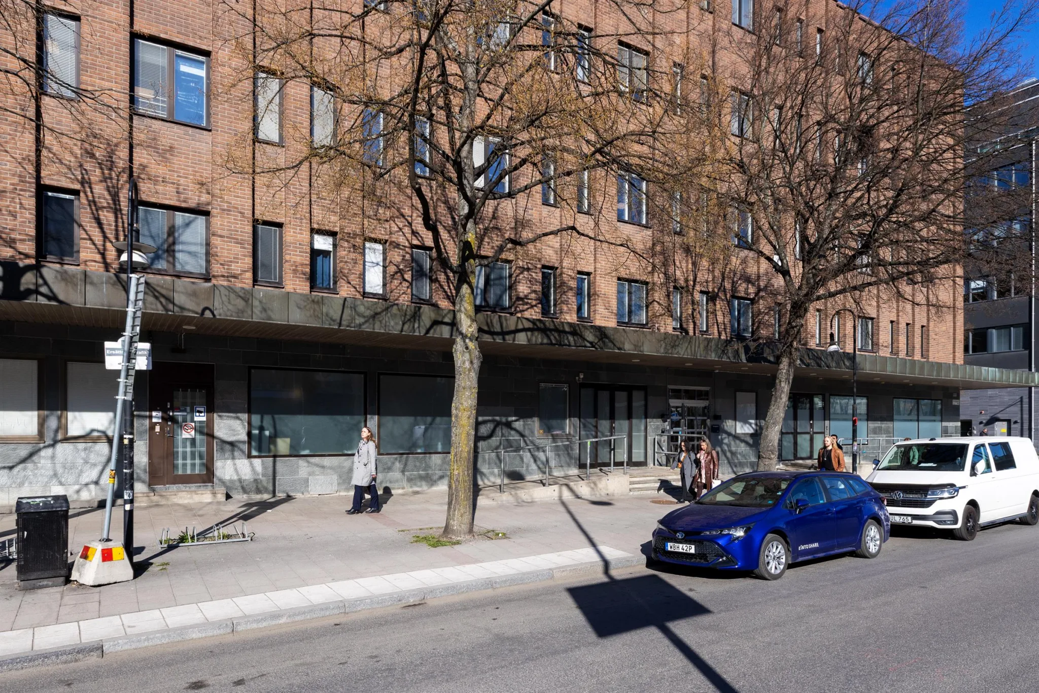 5. Tegelbyggnad med stora fönster och träd framför, en blå bil parkerad på gatan.