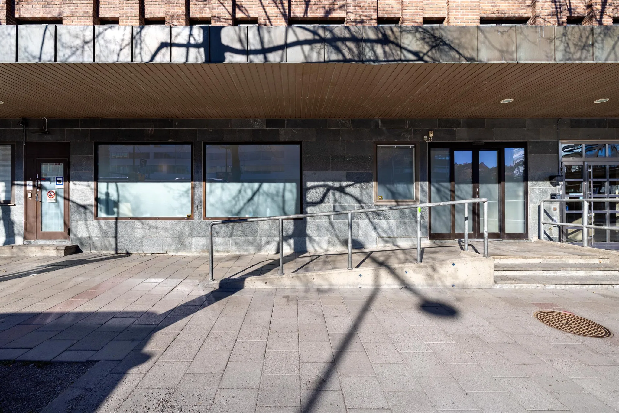 1. Fasad av en byggnad med stora fönster och en ramp framför, med skuggor från träd på marken.