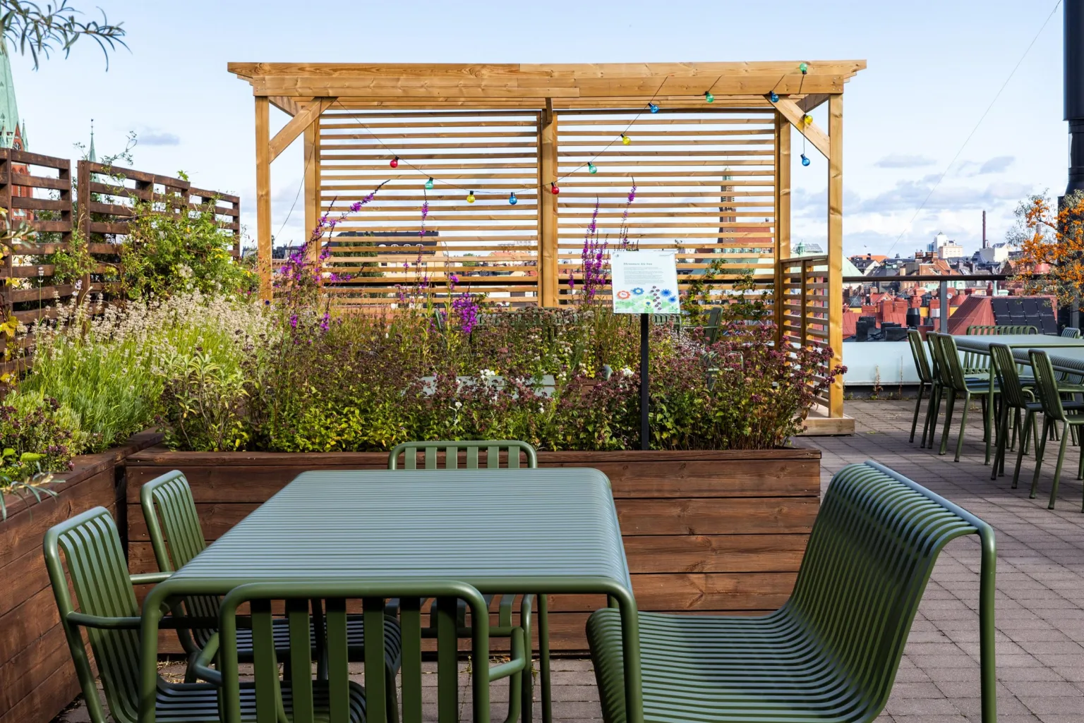 En utomhusterrass med gröna metallmöbler, blomlådor och ett träpergola med färgglada dekorationer.