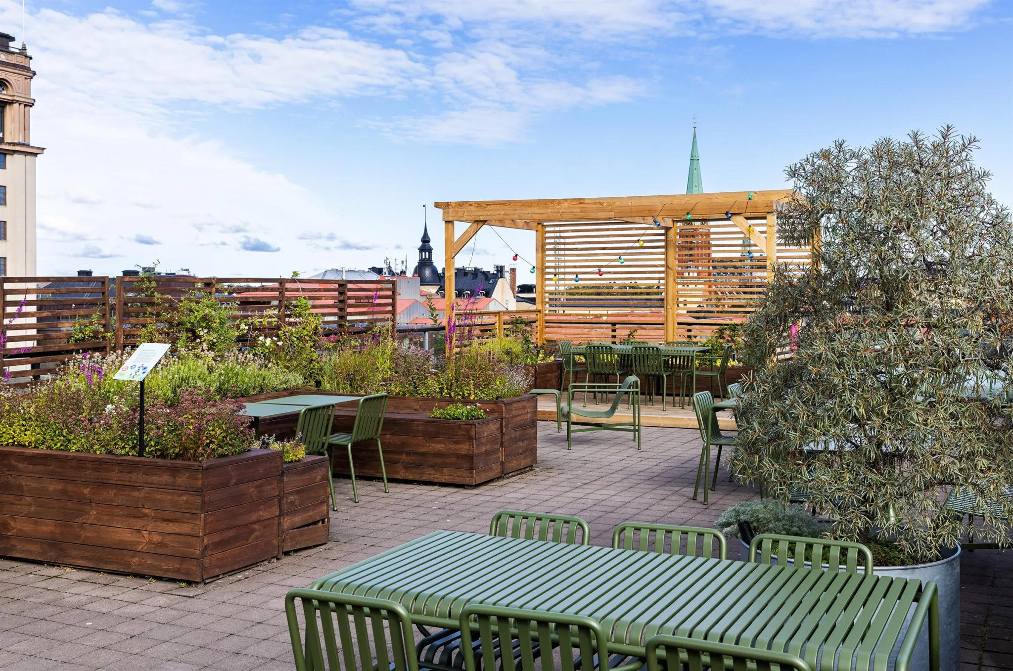 En takterrass med gröna bord, stolar och stora planteringslådor under en blå himmel.