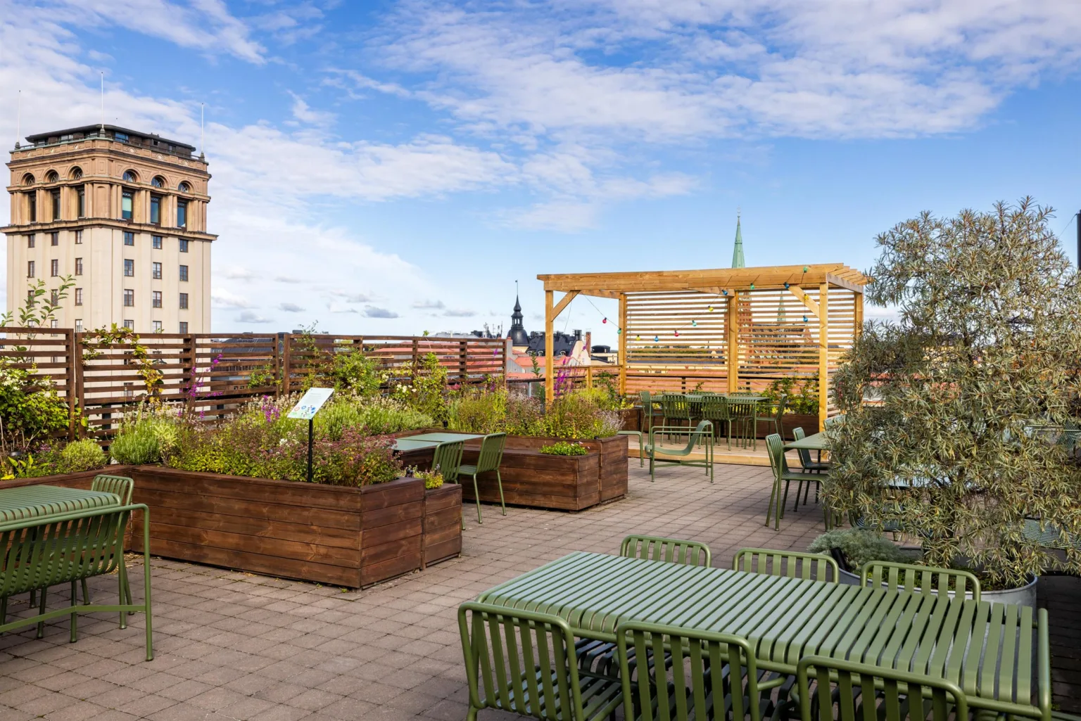 En takterrass med gröna möbler och träplanteringslådor, omgiven av stadsvyer och blå himmel.