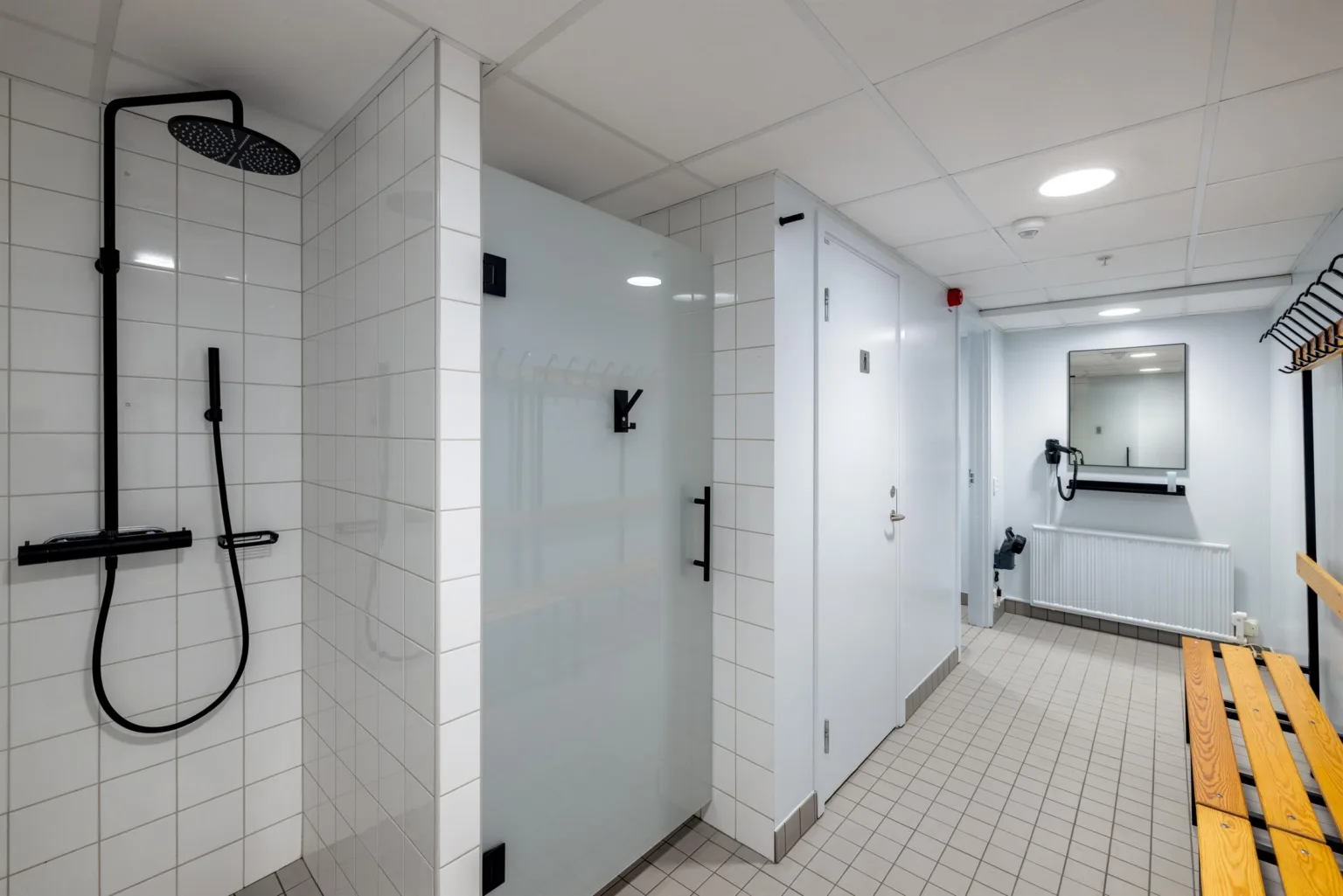 Ett ljust duschutrymme med vita kakelväggar, duschar till vänster och dörrar till duschkabiner.