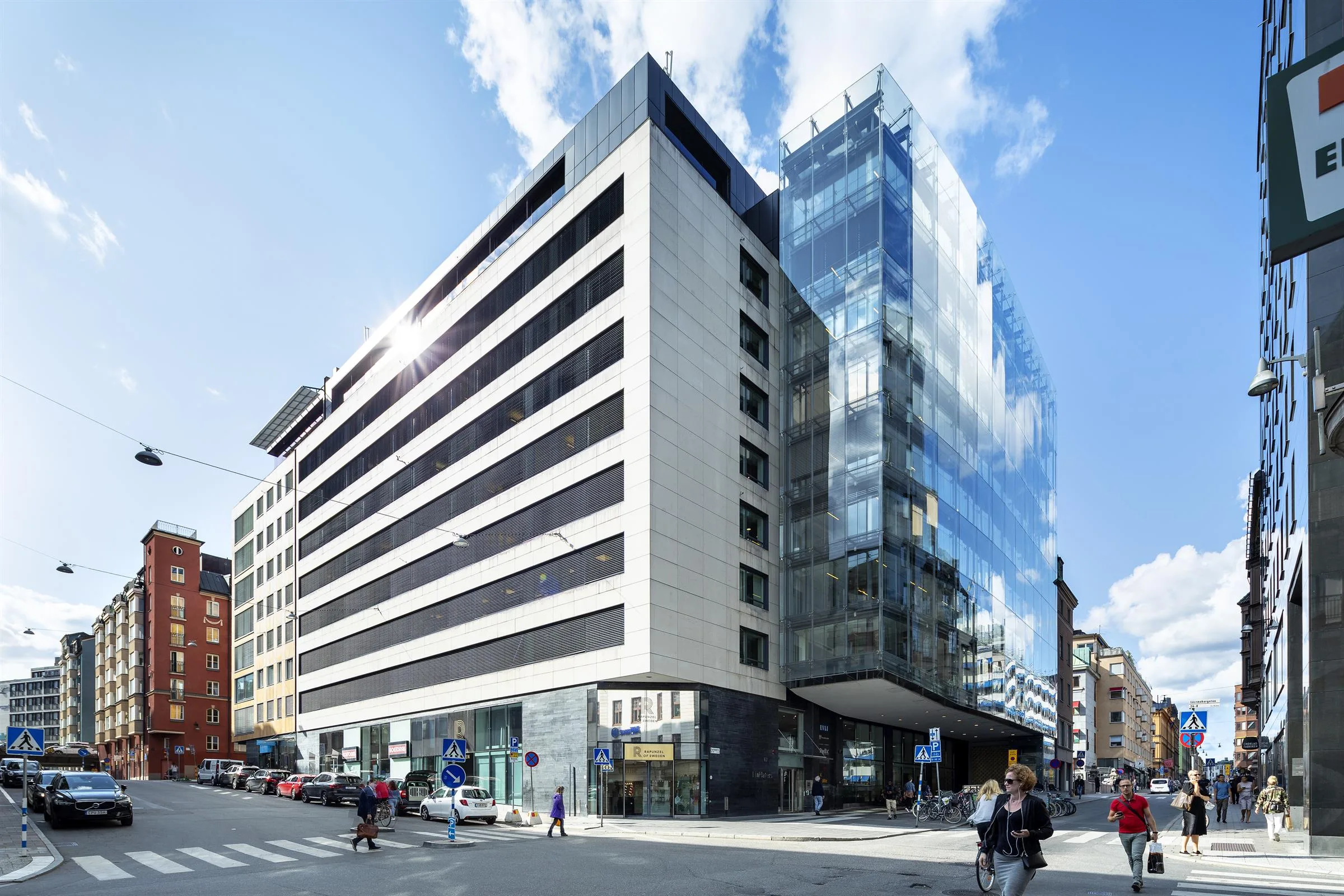 En modern byggnad med glasfasad och vita väggar vid en stadsintersektion, människor går på trottoaren.