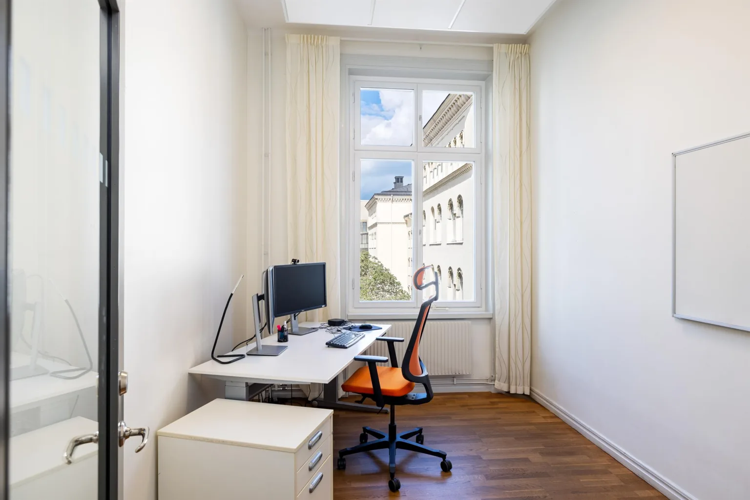 Ett ljust hemmakontor med skrivbord, datorskärm och en orange kontorsstol, samt ett fönster med utsikt över byggnader.