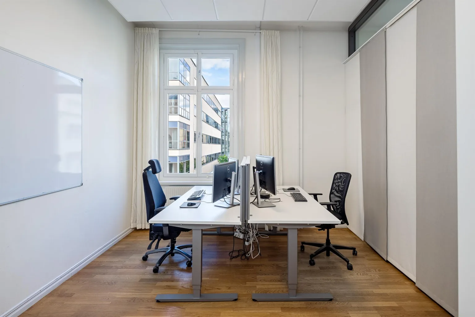 1. Ett ljust kontorsrum med två skrivbord, datorskärmar och fyra svarta kontorsstolar, med ett stort fönster i bakgrunden.
