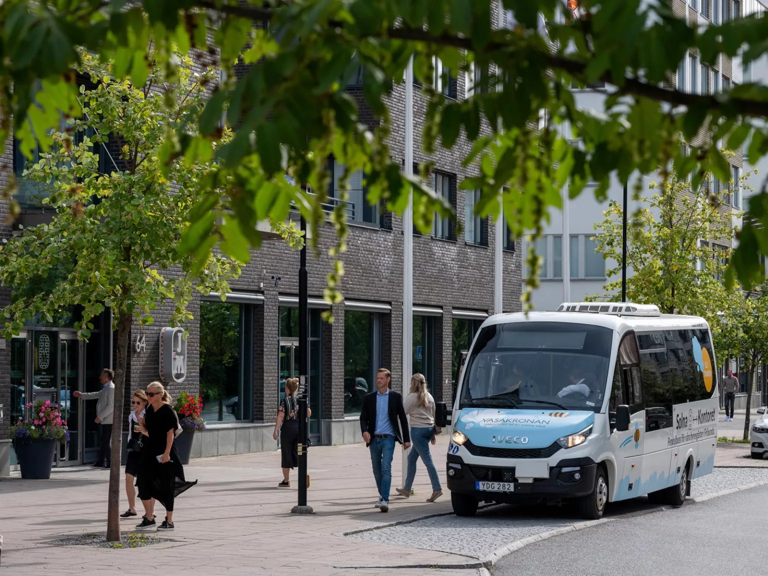 En blå och vit minibuss kör förbi ett område med träd och gångvägar, människor går på trottoaren.