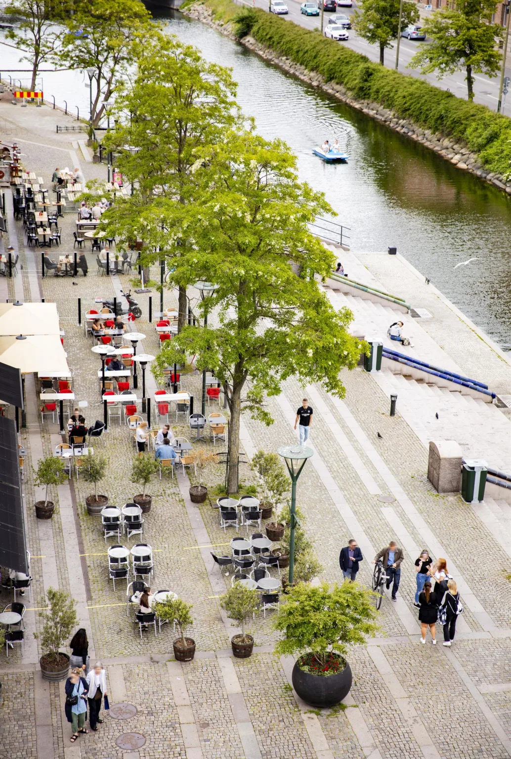 5. Vy över en promenad vid en kanal med uteserveringar, träd och människor som promenerar.