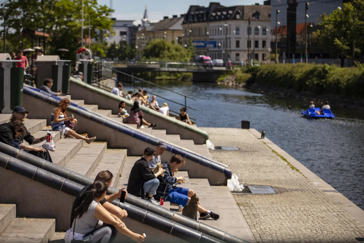 Människor sitter på trappsteg vid en kanal i en stadsmiljö, med grönska och byggnader i bakgrunden.