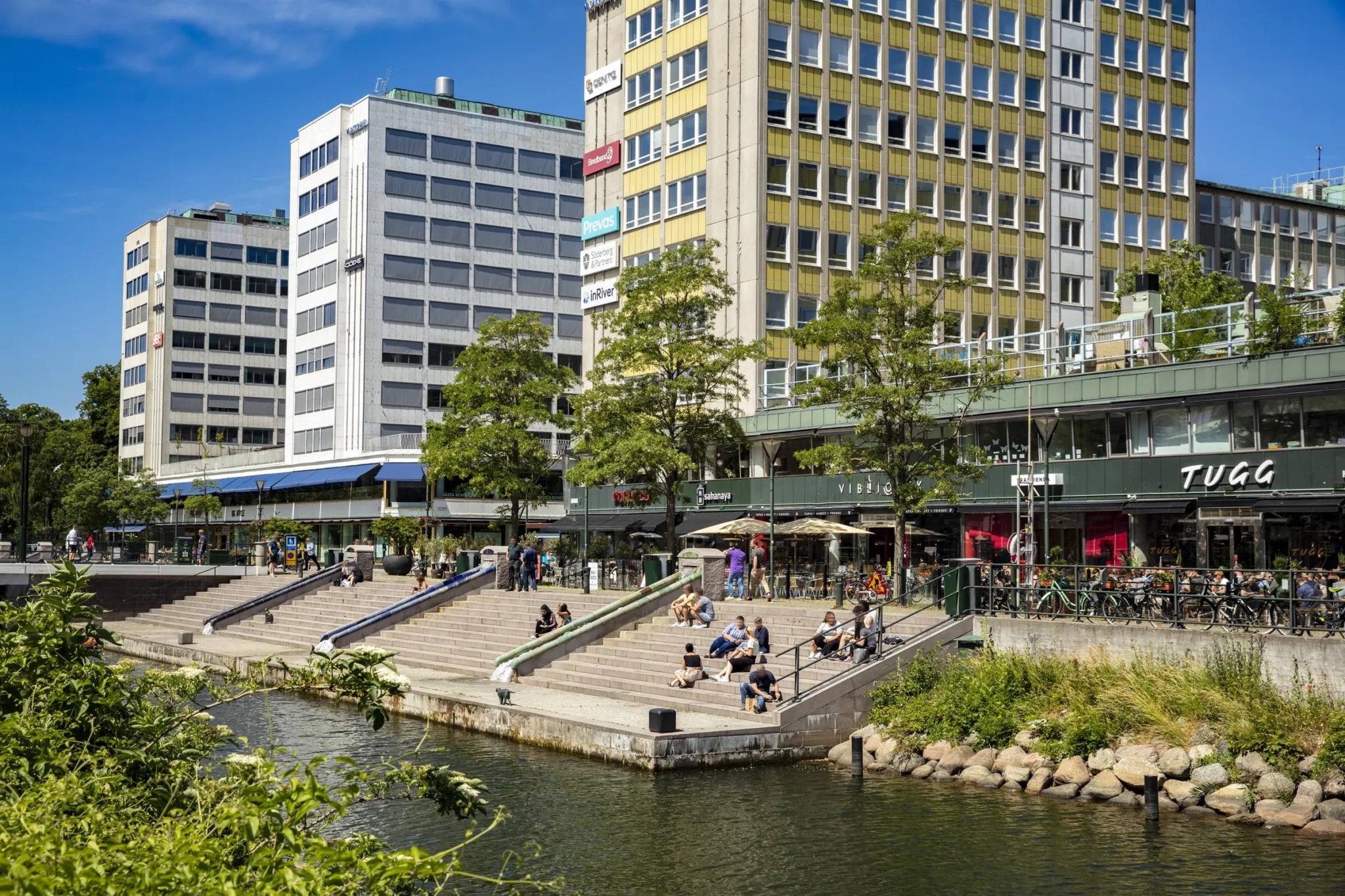 5. En stadsbild med moderna byggnader, grönskande träd, en vattendrag och människor som sitter vid en trappa nära vattnet.