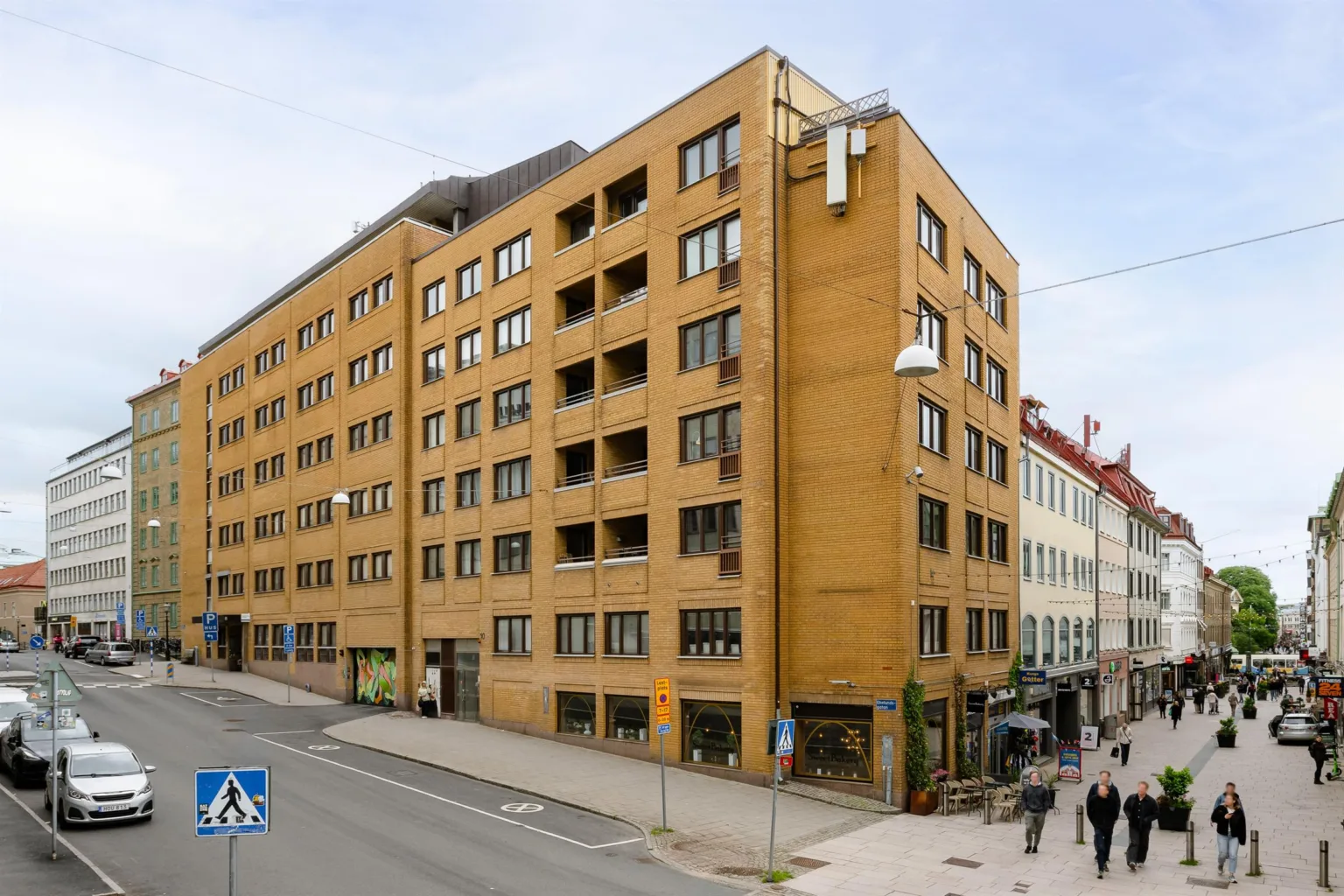 En gul tegelbyggnad med flera våningar, stora fönster och en trottoar med människor utanför.
